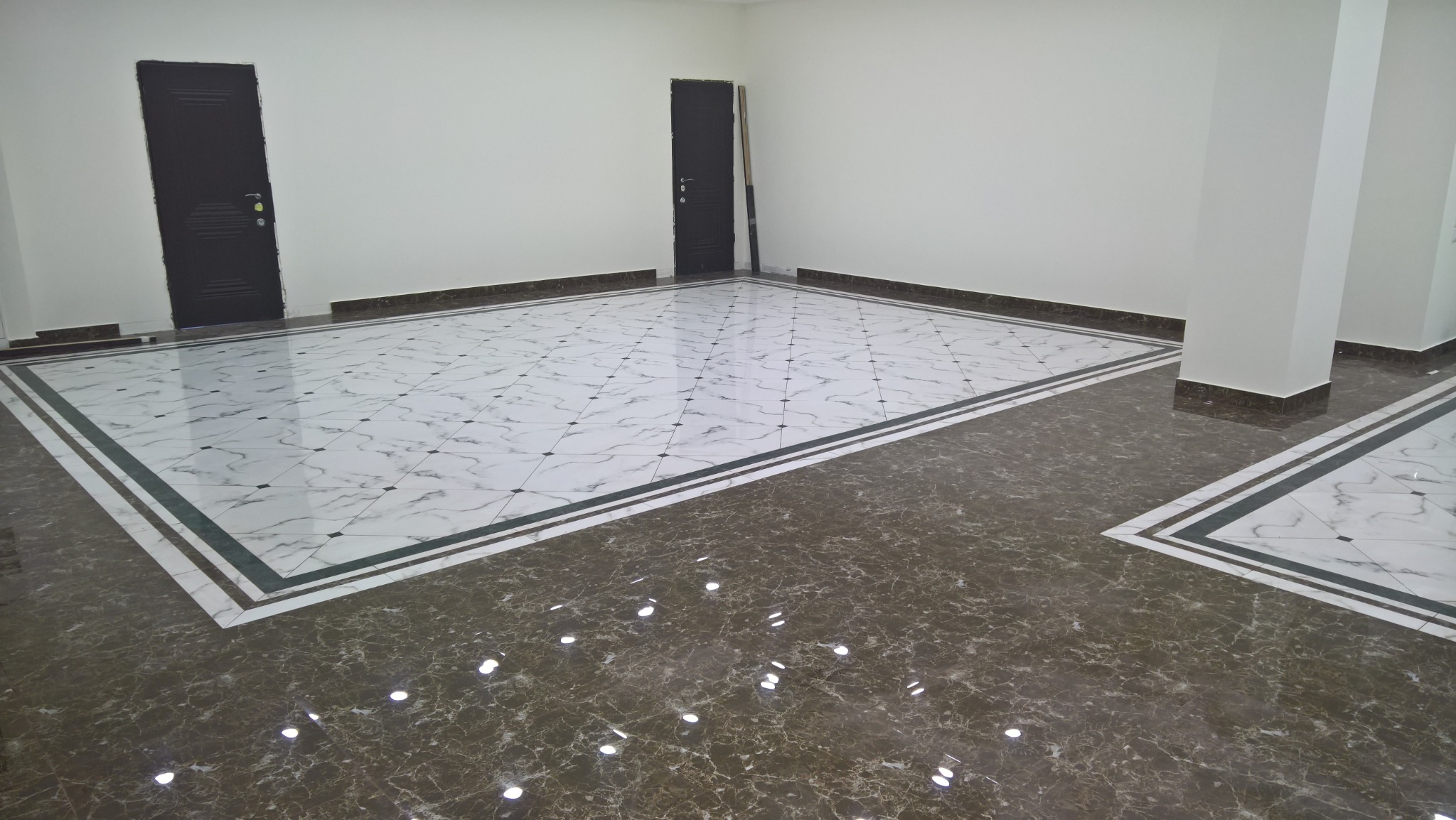 Wykończona podłoga z białych i brązowych marmurowych płytek z ciemnymi listwami przypodłogowymi i dwiema czarnymi, zamkniętymi drzwiami w pustym pomieszczeniu.
