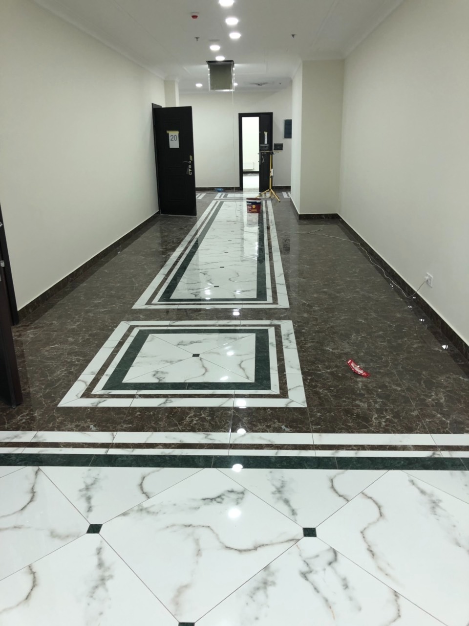 Korytarz z nową podłogą wyłożoną brązowymi i białymi płytkami imitującymi marmur, z dekoracyjnymi wstawkami i listwami, widok na drzwi z numerem 20 i dalszą część korytarza.