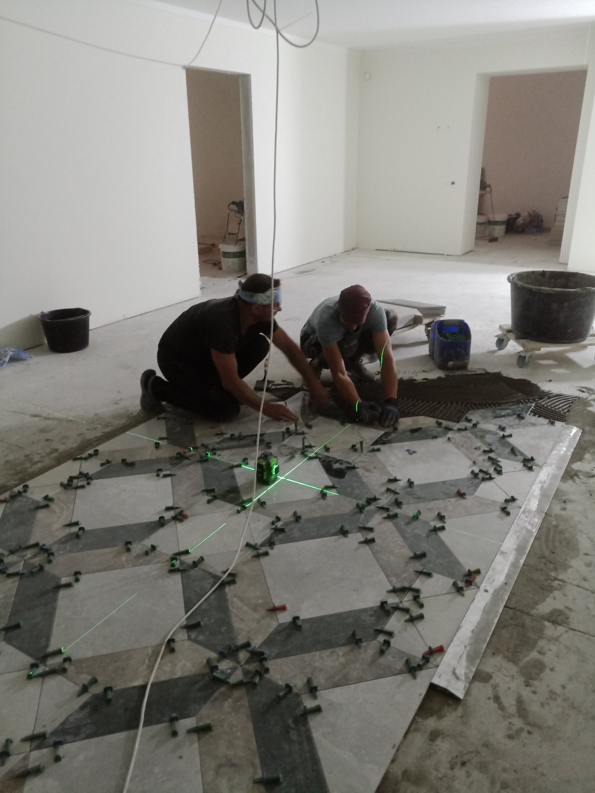 Dwóch mężczyzn układa wzorzystą podłogę z płytek, używając poziomicy laserowej i klipsów dystansowych w surowym wnętrzu.