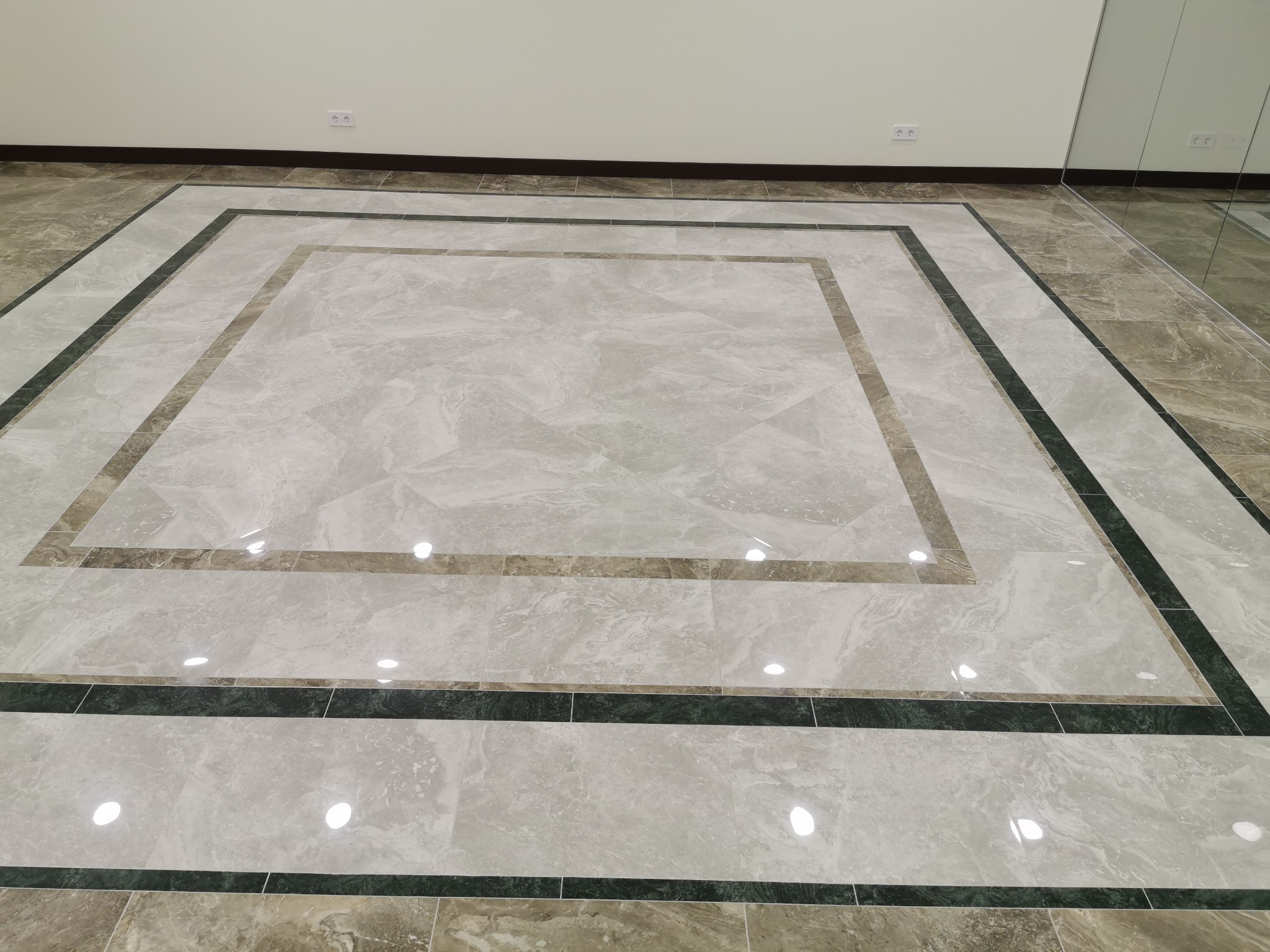 Wzorzysta podłoga z jasnych, marmurkowych płytek z ciemnozielonym i beżowym obramowaniem, widoczne gniazdka elektryczne na ścianie i fragment lustrzanej ściany.