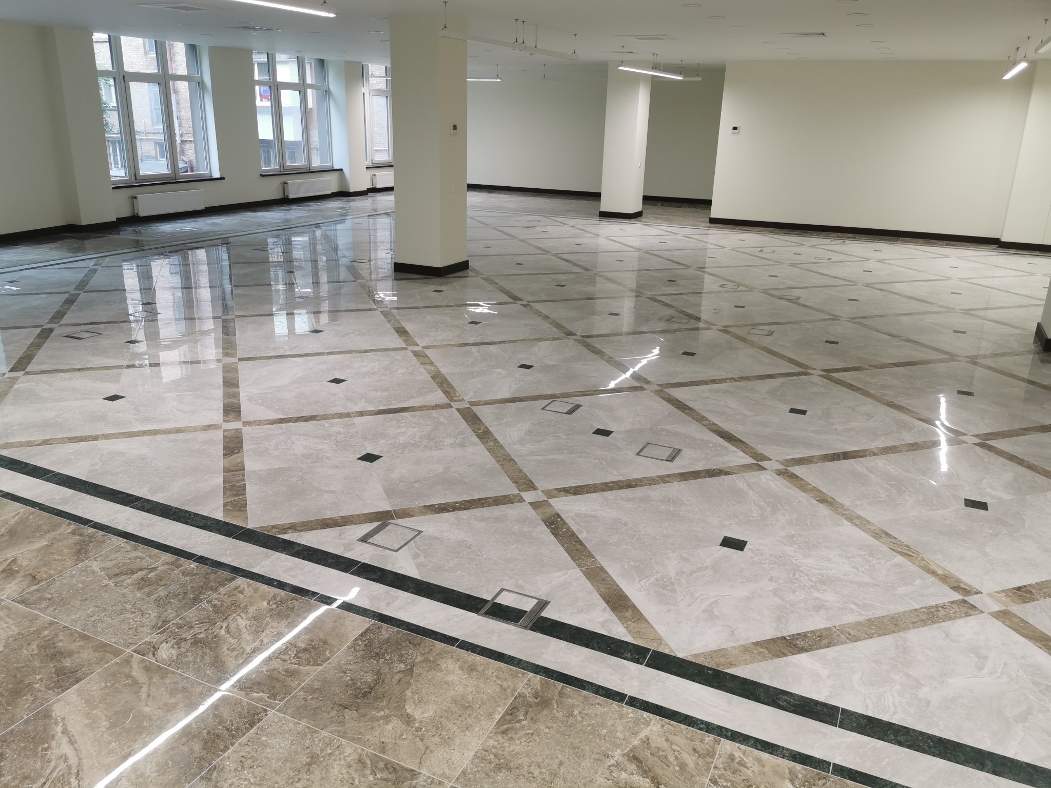 Wykończona podłoga biurowa z jasnoszarych, kwadratowych płytek marmurowych z ciemniejszymi, brązowymi wstawkami i czarnymi kwadratami, widoczne słupy i okna.