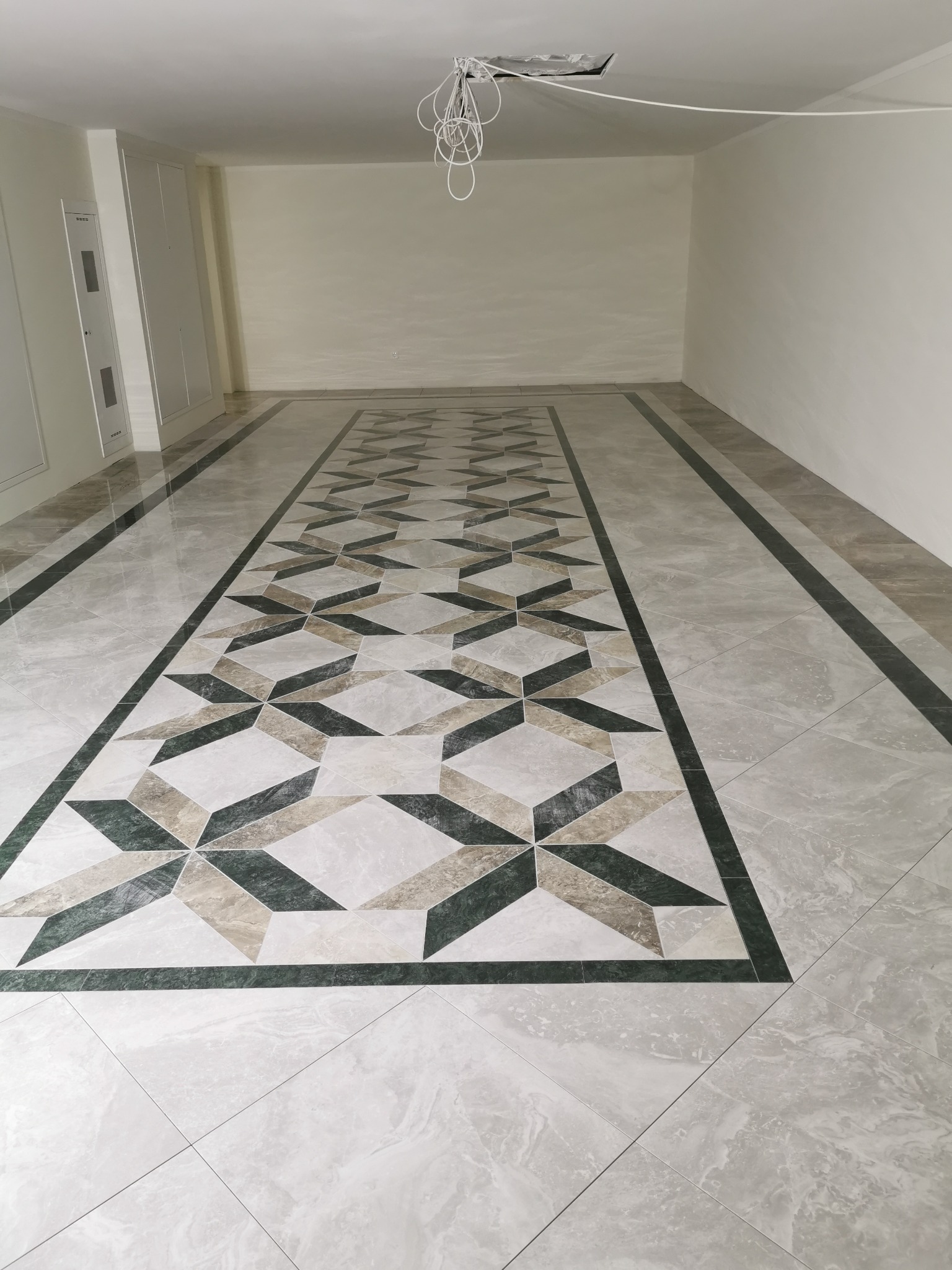 Wnętrze z nową, wzorzystą podłogą z płytek w odcieniach szarości, beżu i zieleni, ułożoną w geometryczny deseń na środku pomieszczenia, obramowaną ciemnozielonym pasem.