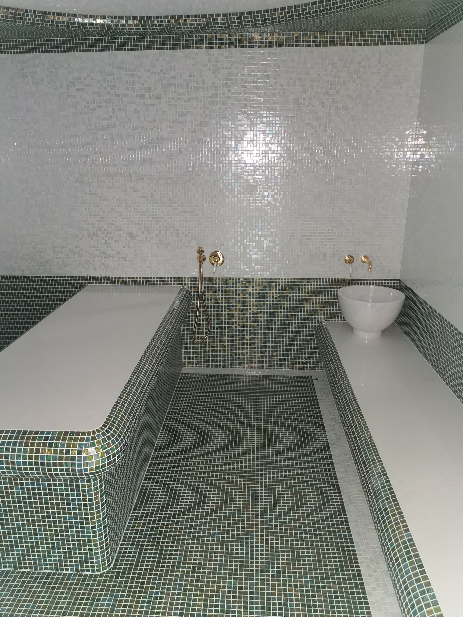 Wnętrze łaźni wykończone mozaiką w odcieniach bieli, zieleni i złota, z umywalką w kształcie misy i złotą armaturą.