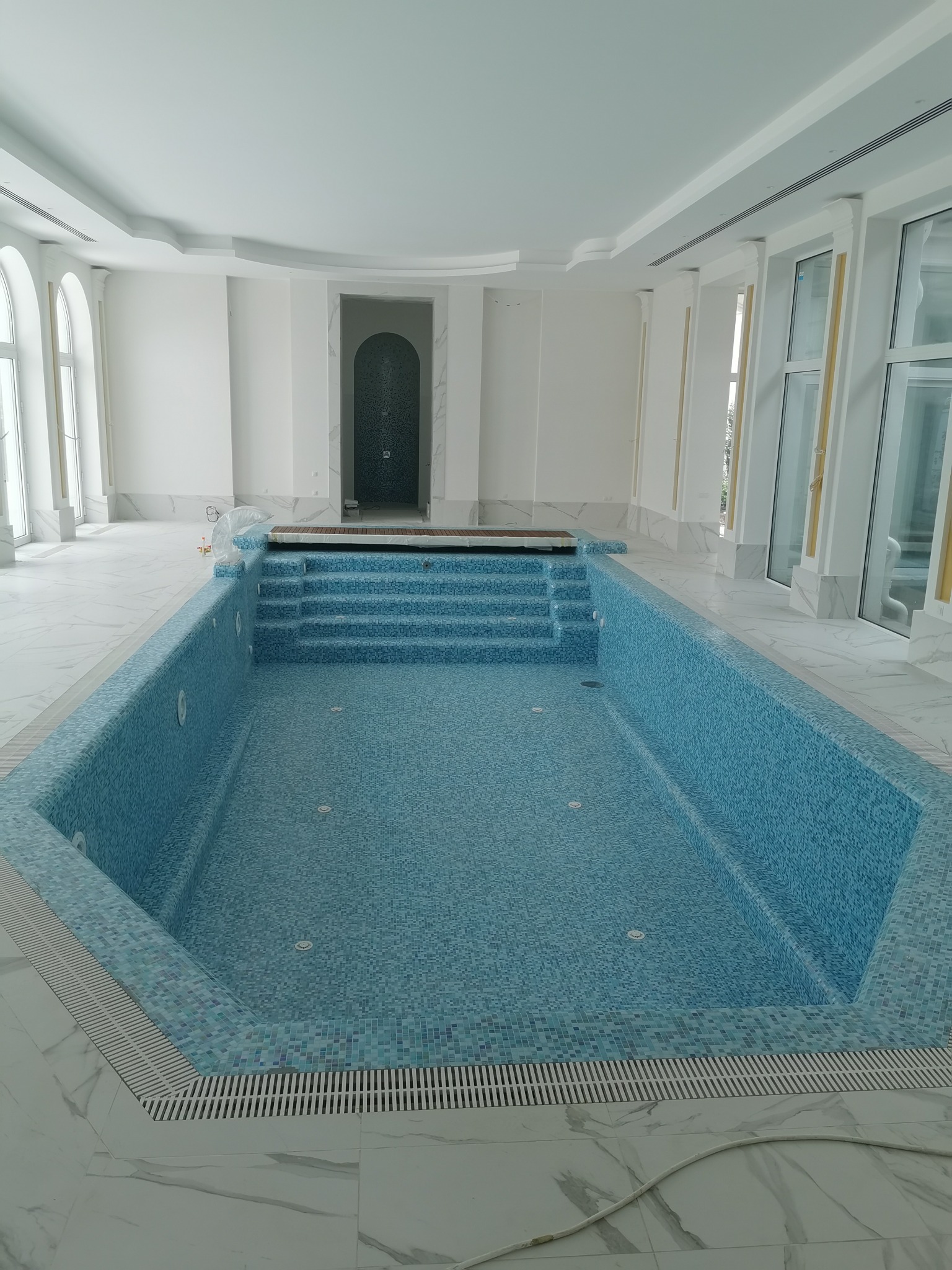 Wnętrze basenu wyłożonego jasnoniebieską mozaiką, z białymi marmurowymi podłogami i ścianami, widoczne schody wejściowe do basenu i kratki odpływowe.