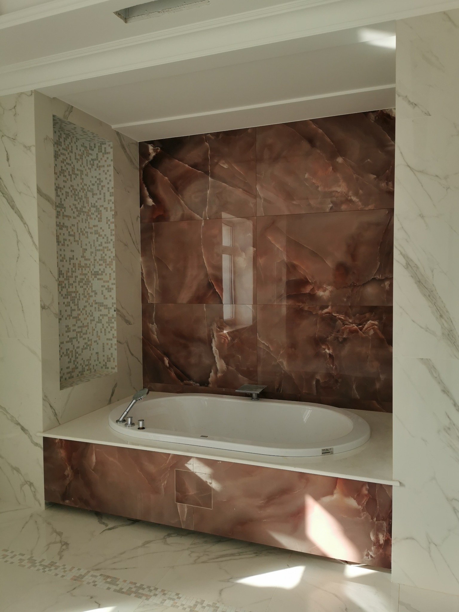 Nowoczesna łazienka z owalną wanną obudowaną panelami imitującymi marmur, ściany wyłożone płytkami marmurowymi w odcieniach brązu i bieli, dekoracyjna wnęka z mozaiką.