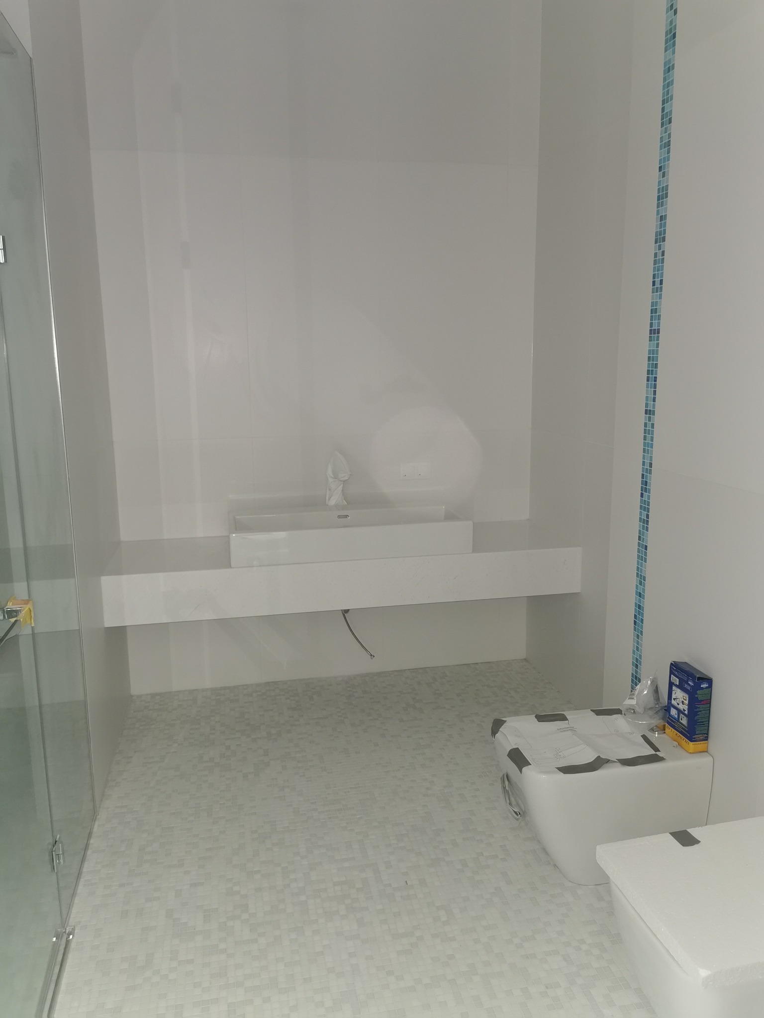 Nowoczesna, minimalistyczna łazienka z białymi płytkami mozaikowymi na podłodze, wisząca umywalka z blatem i szklana kabina prysznicowa, toaleta przygotowana do montażu z przyklejoną instrukcją.