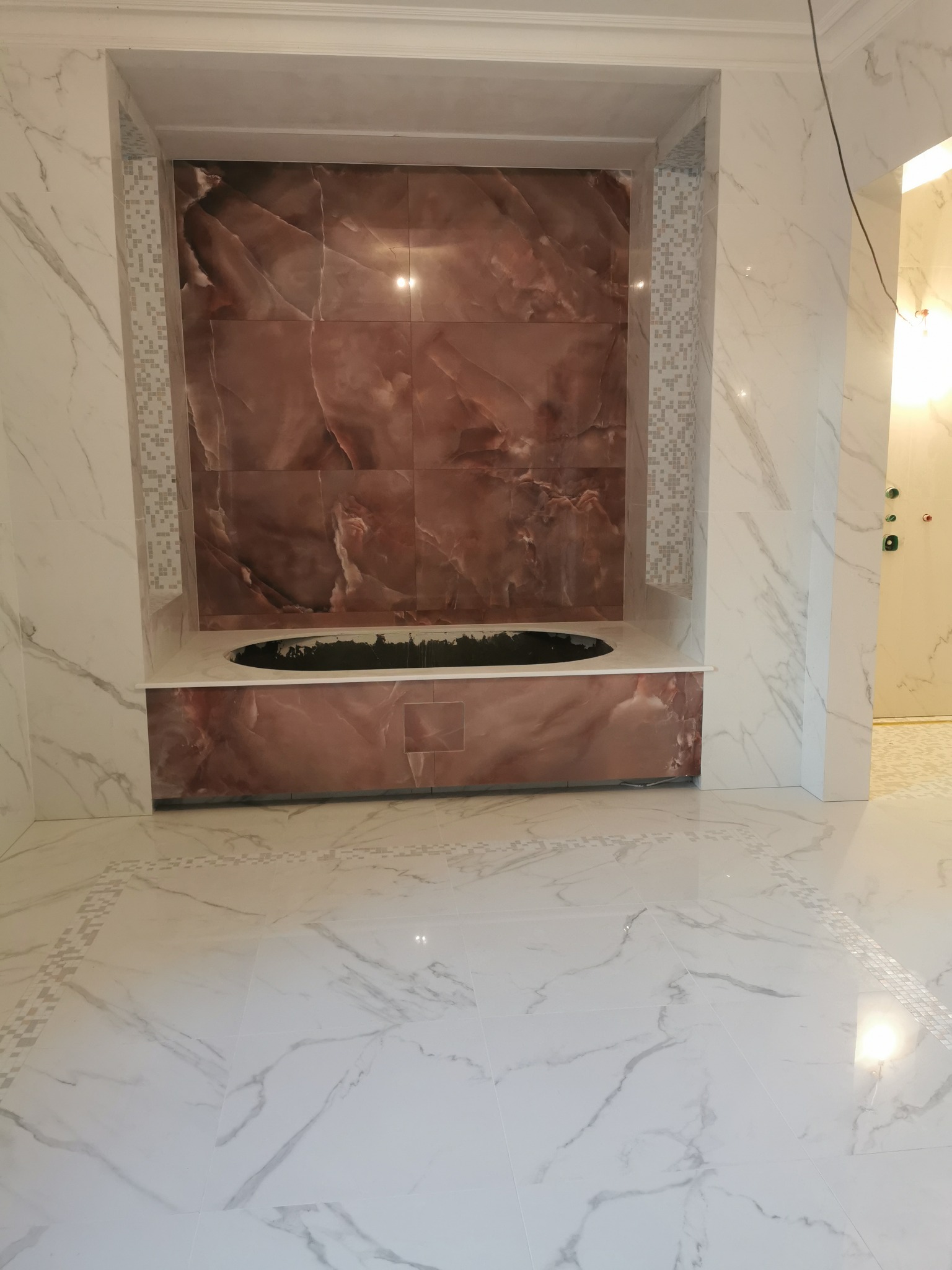 Wykończona wnęka z brązowymi płytkami imitującymi marmur nad wanną, otoczona białymi płytkami z szarymi żyłami, mozaika na ścianach wnęki i na podłodze.