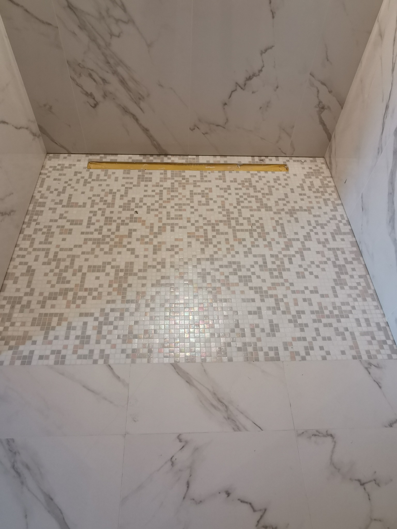 Wykończony brodzik prysznicowy z odpływem liniowym, wyłożony mozaiką z jasnych płytek i obłożony marmurową imitacją na ścianach.