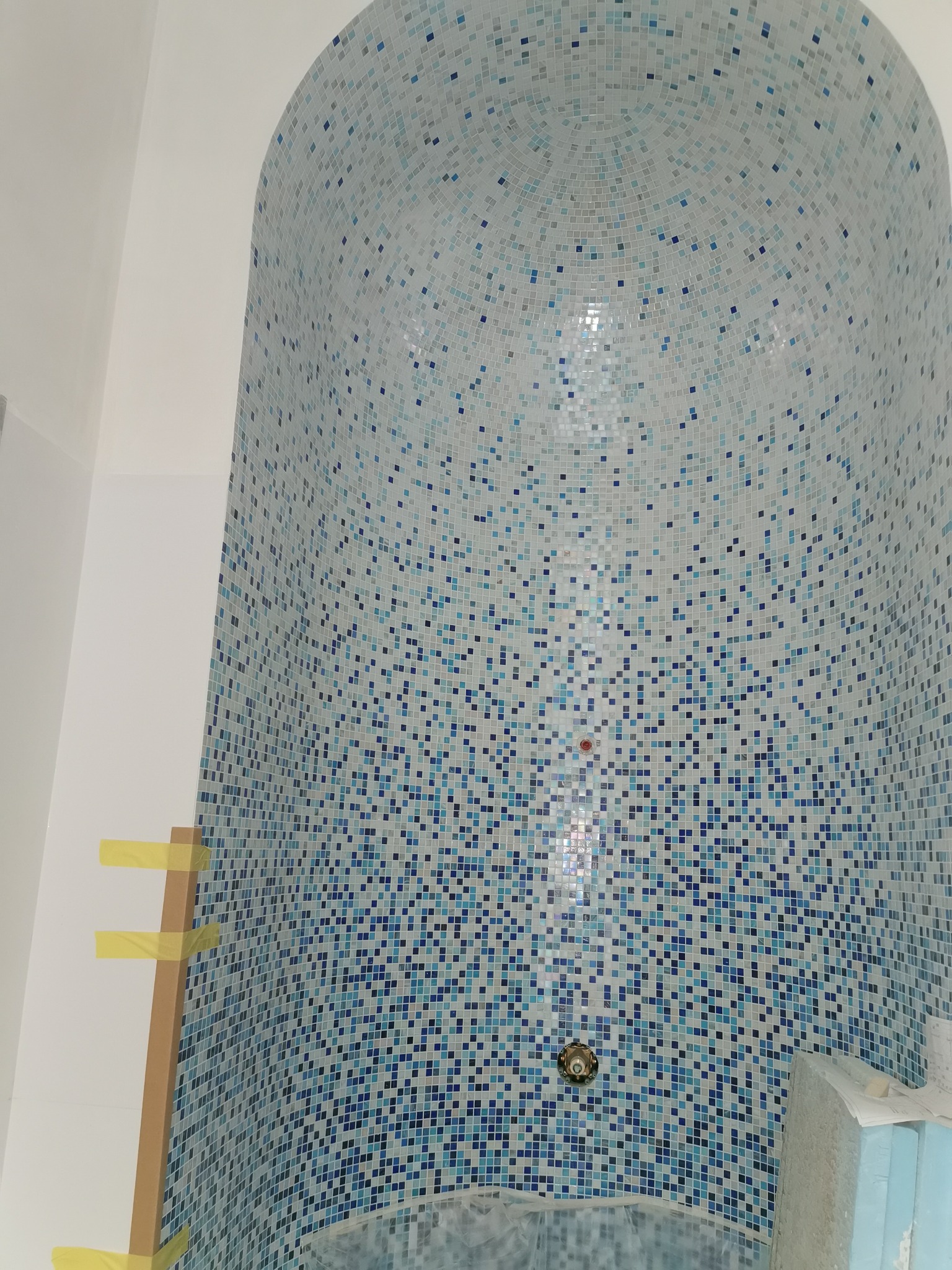Półokrągła wnęka w łazience wyłożona mozaiką z drobnych kwadratowych płytek w odcieniach błękitu i szarości, widoczne zabezpieczenia taśmą malarską i fragment instalacji wodnej.