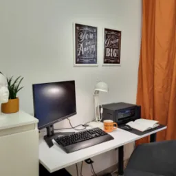 Domowe biuro z białym biurkiem, monitorem, klawiaturą, lampką, drukarką, notatnikiem, pomarańczowym kubkiem Bistro, słuchawkami na manekinie głowy i motywującymi plakatami na ścianie.