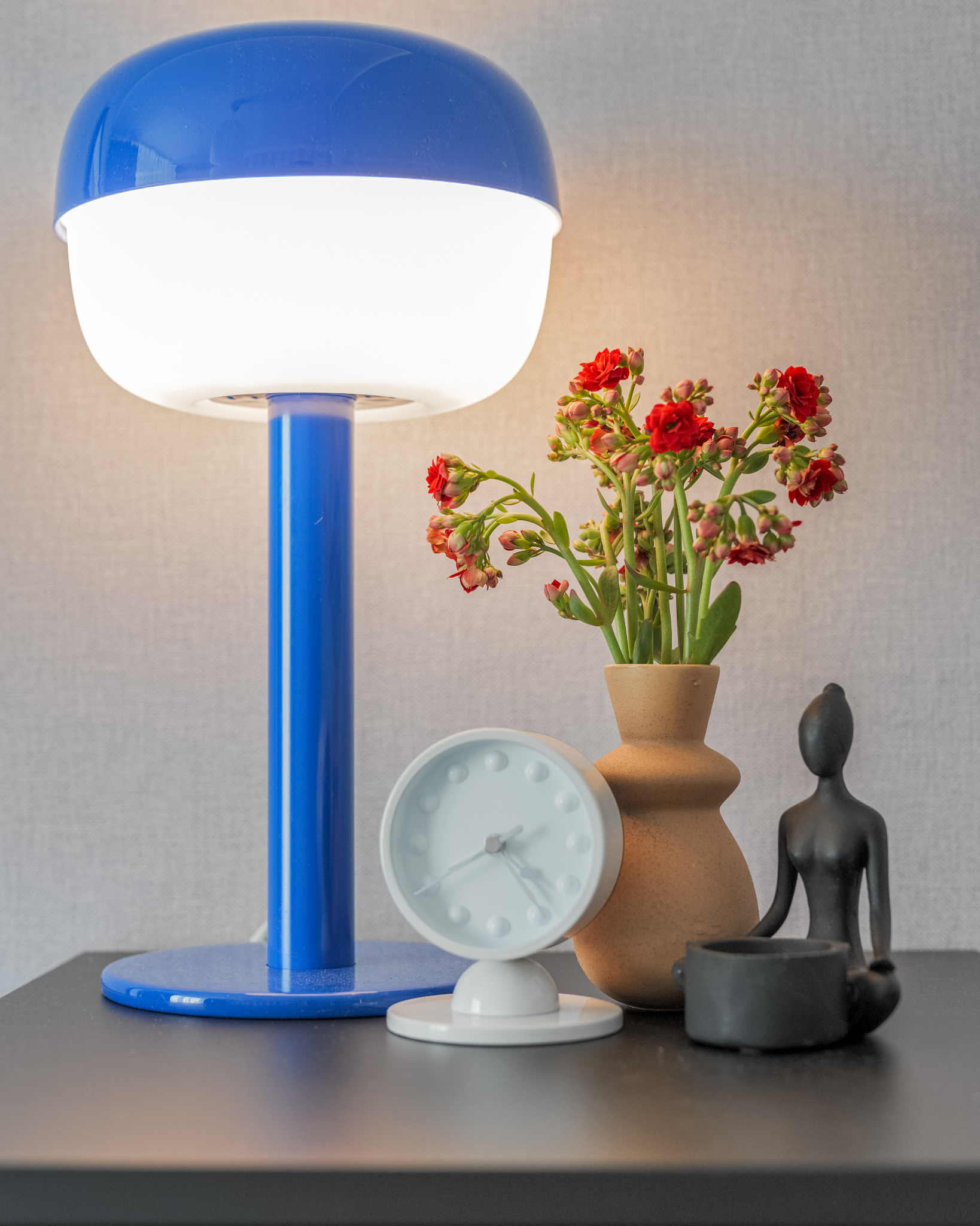 Nastrojowe ujęcie detali: niebieska lampka nocna, zegar z wypukłymi oznaczeniami godzin, wazon z czerwonymi kwiatami oraz minimalistyczna figurka na czarnym stoliku.