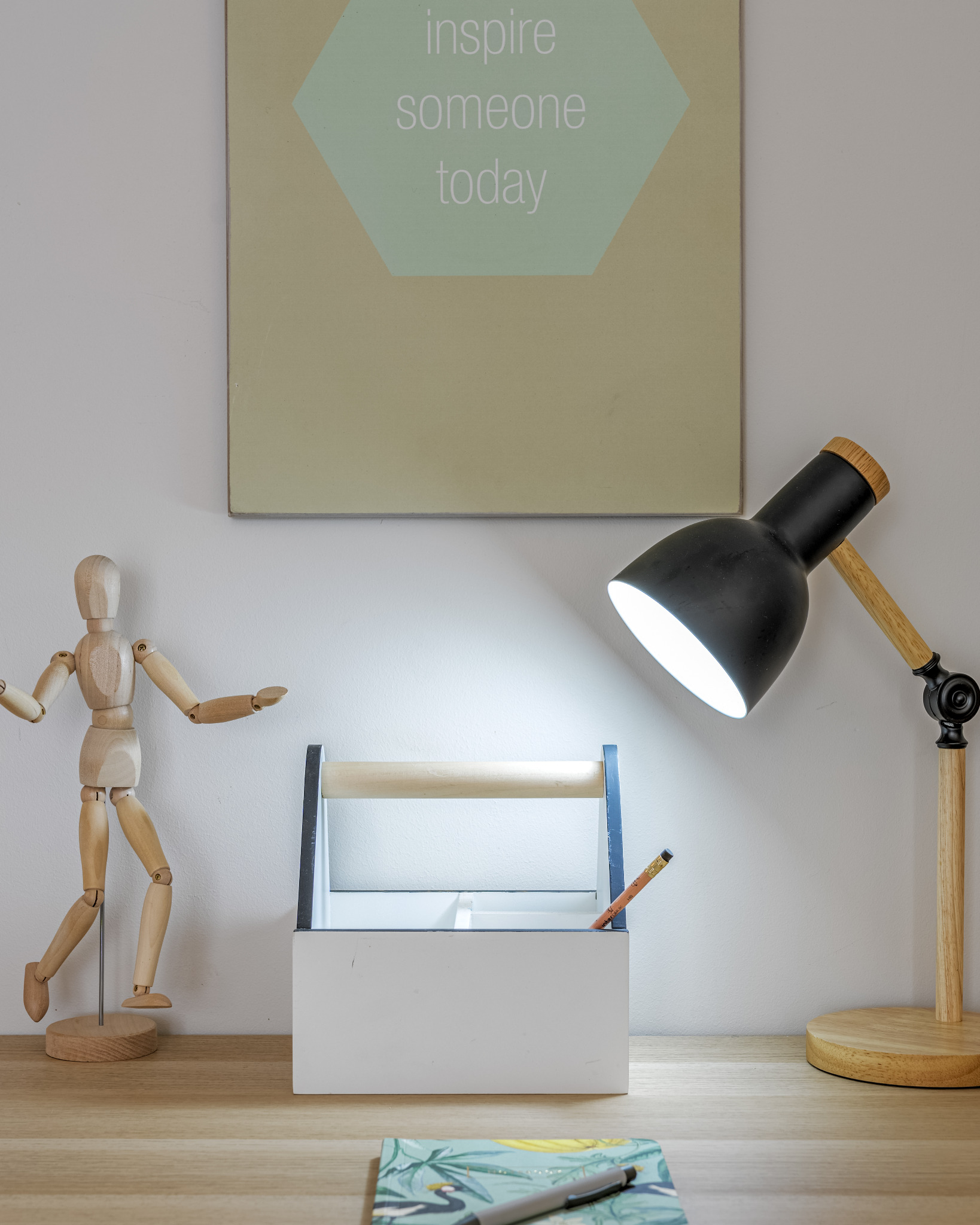 Drewniany manekin, lampka nocna z drewnianymi elementami, biały organizer z uchwytem i napis 'inspire someone today' na ścianie, tworzą minimalistyczną kompozycję na biurku.