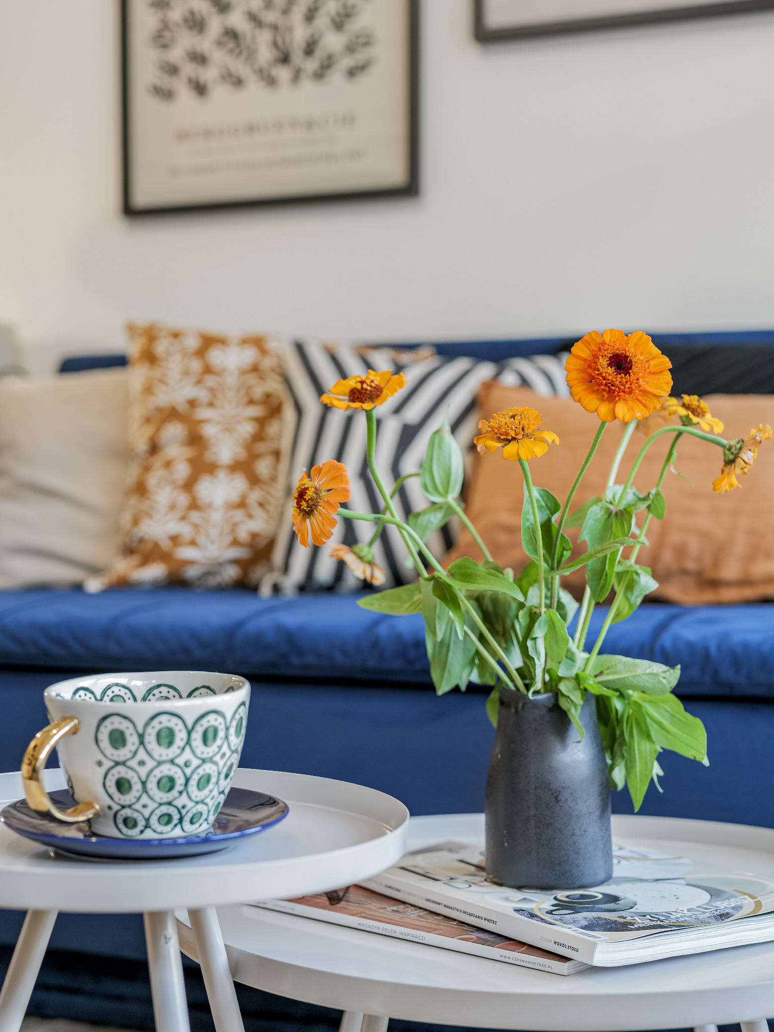 Aranżacja salonu z niebieską sofą, stolikiem kawowym z wazonem z pomarańczowymi kwiatami i filiżanką z zielonym wzorem.