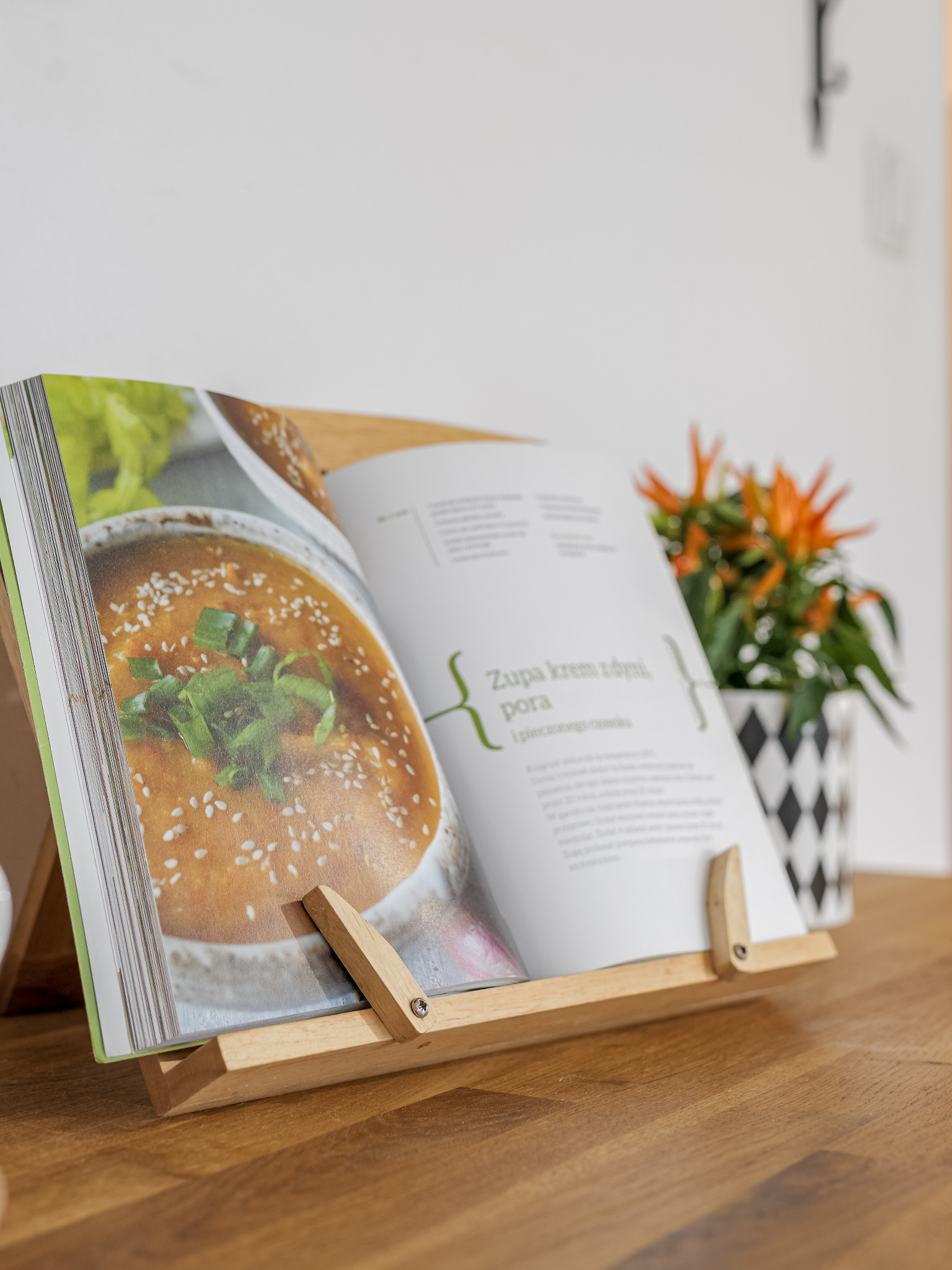 Otwarta książka kucharska na drewnianym stojaku, prezentująca zdjęcie kremowej zupy z posiekanym szczypiorkiem i sezamem, obok doniczka z pomarańczowymi kwiatami w czarno-białej geometrycznej...