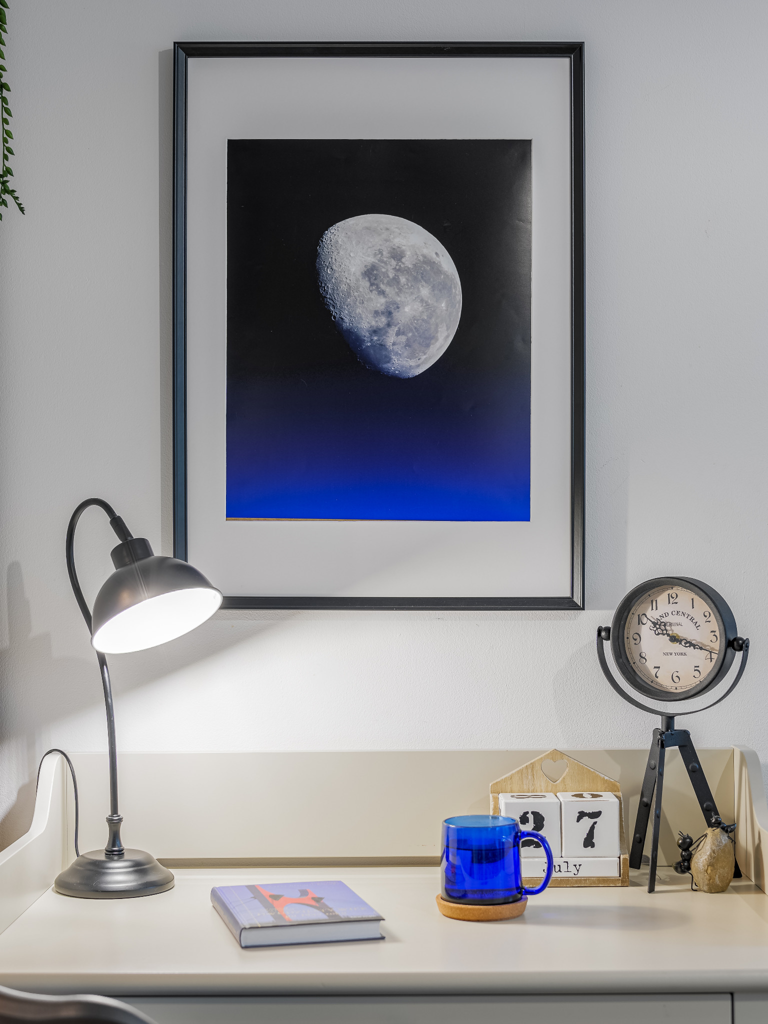 Białe biurko z lampką, książką, niebieskim kubkiem na podkładce, zegarem na trójnogu i kalendarzem wskazującym 27 lipca, na ścianie wisi oprawiony plakat z księżycem.