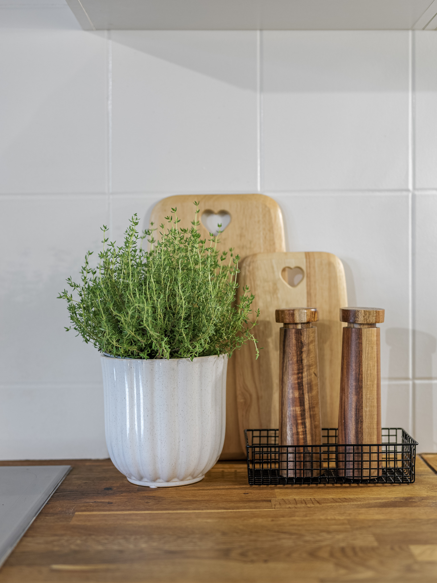 Dekoracja kuchenna: doniczka z tymiankiem, drewniane deski do krojenia z wyciętymi sercami i drewniane młynki do przypraw w czarnym metalowym koszyku na drewnianym blacie na tle białych płytek.