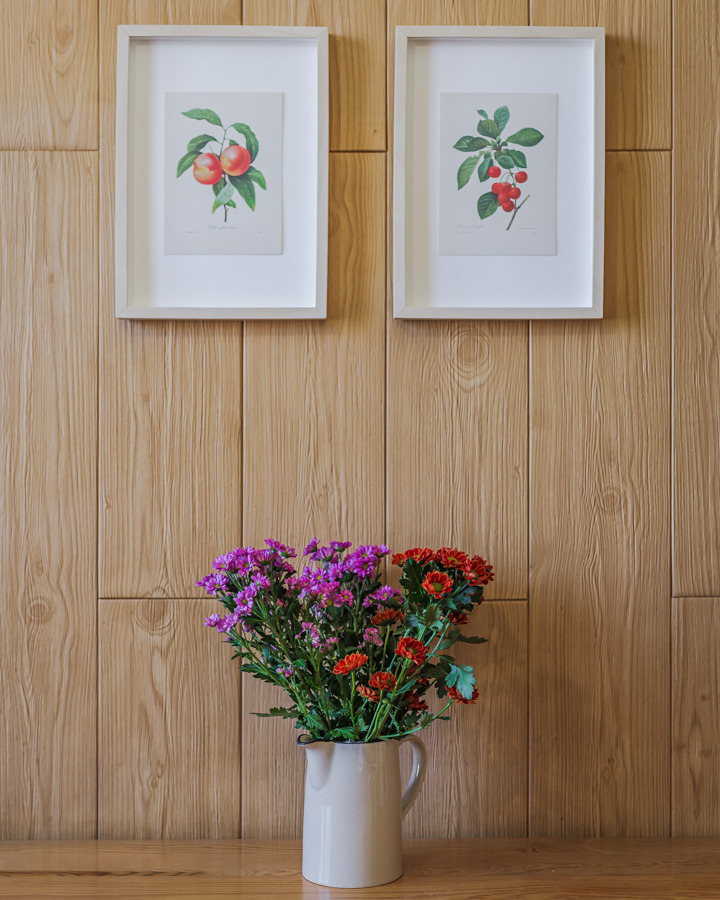 Bukiet fioletowych i czerwonych chryzantem w kremowym dzbanku, wiszące nad nim dwie oprawione grafiki botaniczne z motywem owoców na drewnianej ścianie z paneli.