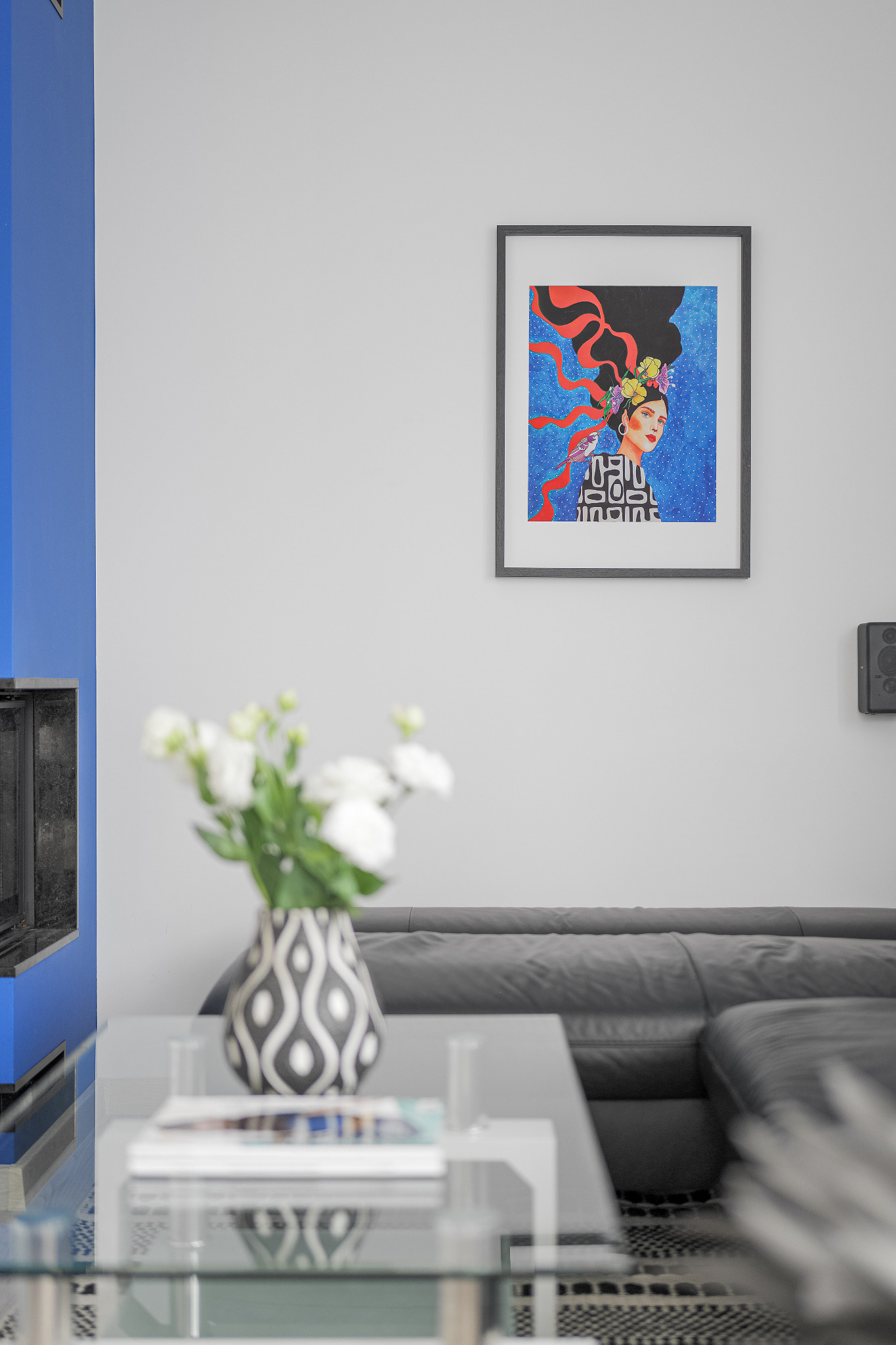 Wnętrze salonu z nowoczesnym kominkiem w niebieskiej obudowie, szarą sofą i szklanym stolikiem kawowym z wazonem białych kwiatów, na ścianie wisi oprawiony portret w stylu inspirowanym Fridą Kahlo.