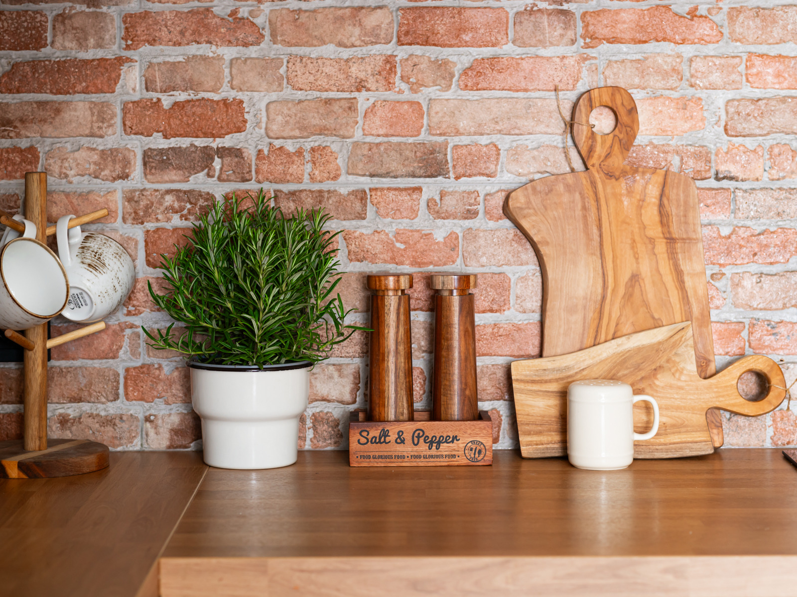Ustawienie na drewnianym blacie: zioła w białej doniczce, drewniane młynki do soli i pieprzu, deski do krojenia z jasnego drewna oraz biały kubek, na tle ściany z czerwonej cegły.