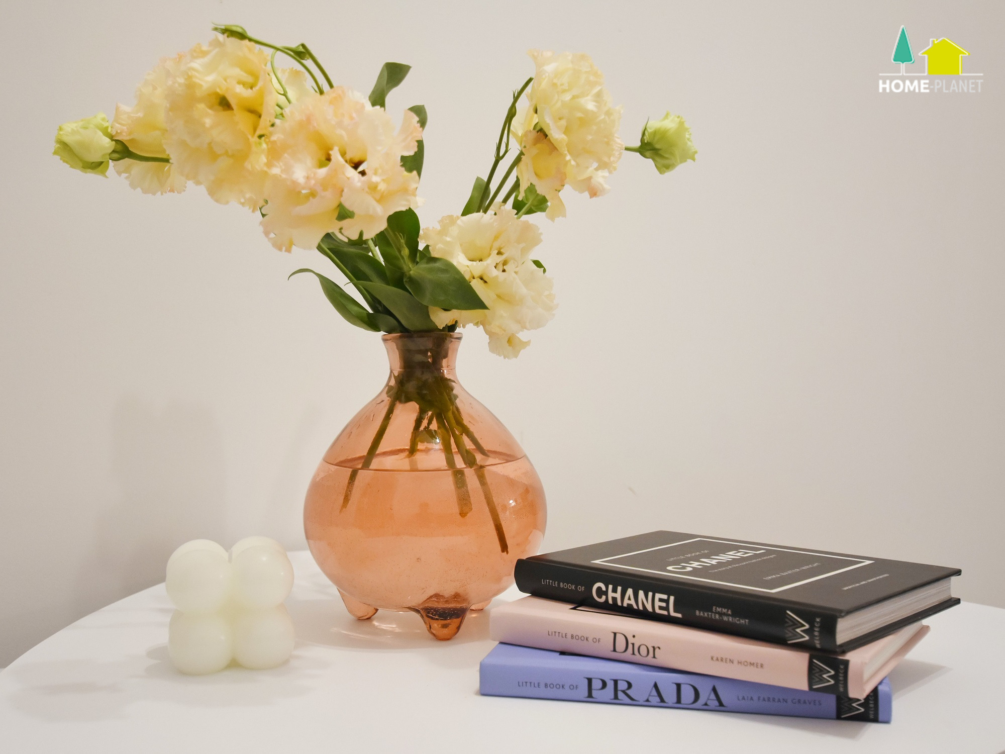 Dekoracyjny bukiet z kremowych kwiatów w szklanym wazonie na nóżkach, obok biała świeczka w kształcie kuli i stos książek o modzie z okładkami Chanel, Dior i Prada, na białym blacie.