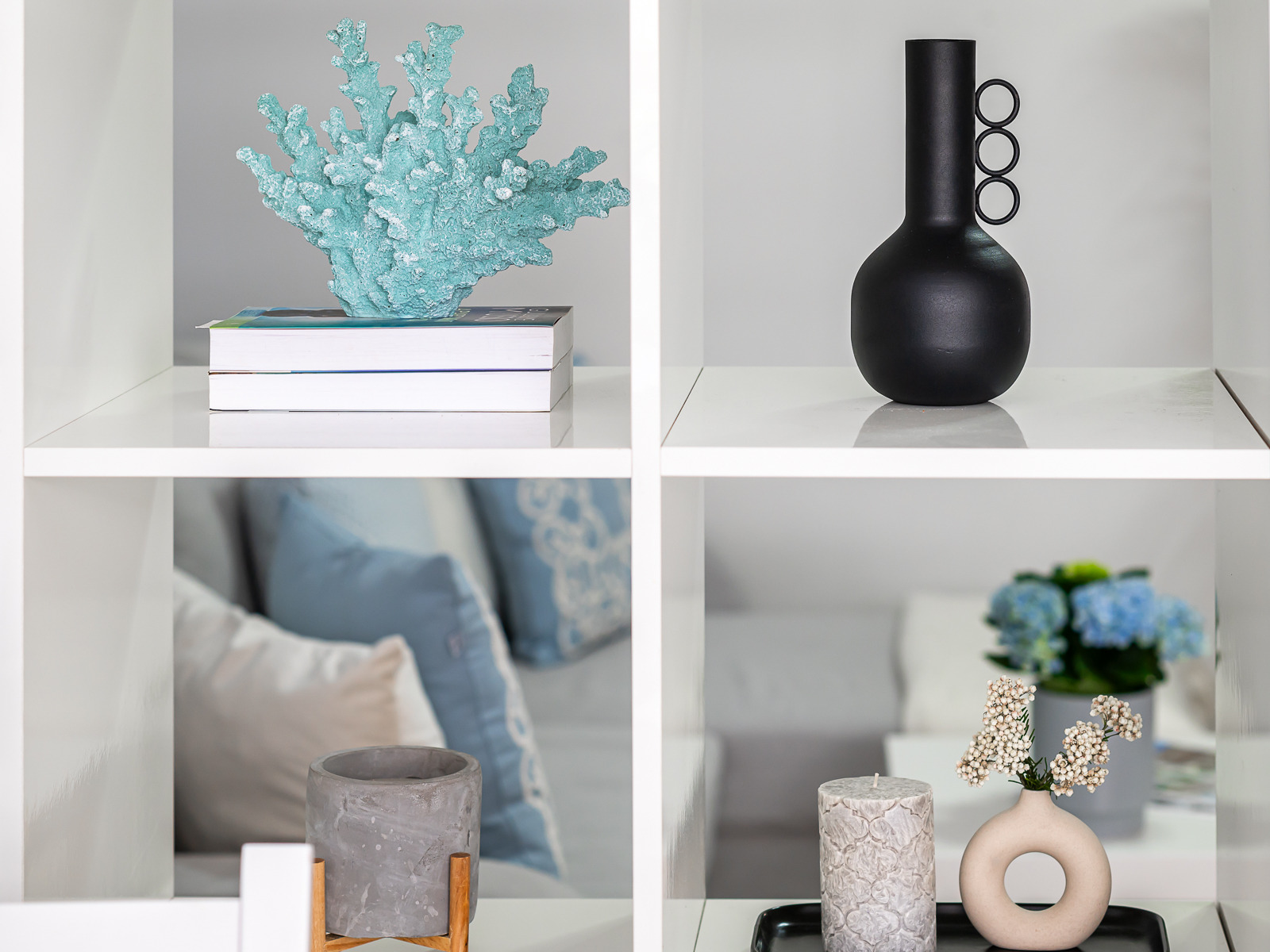 Dekoracyjne przedmioty na białych półkach: turkusowy koral na książkach, czarny wazon z trzema okręgami, betonowa doniczka na drewnianym stojaku, świeca i wazon z białymi kwiatami na czarnej tacy.
