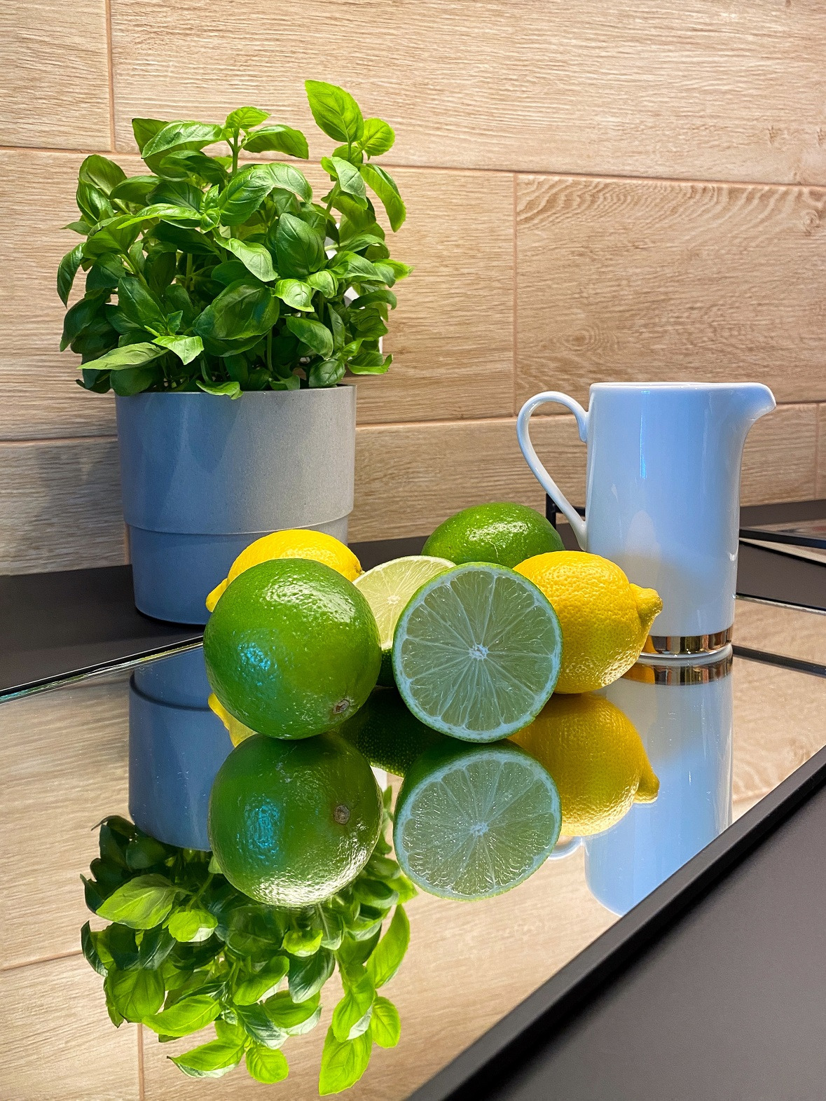 Aranżacja kuchni w Ożarowie Mazowieckim z bazylią w szarej doniczce, cytrynami, limonkami i białym dzbankiem z lustrzanym odbiciem na blacie.