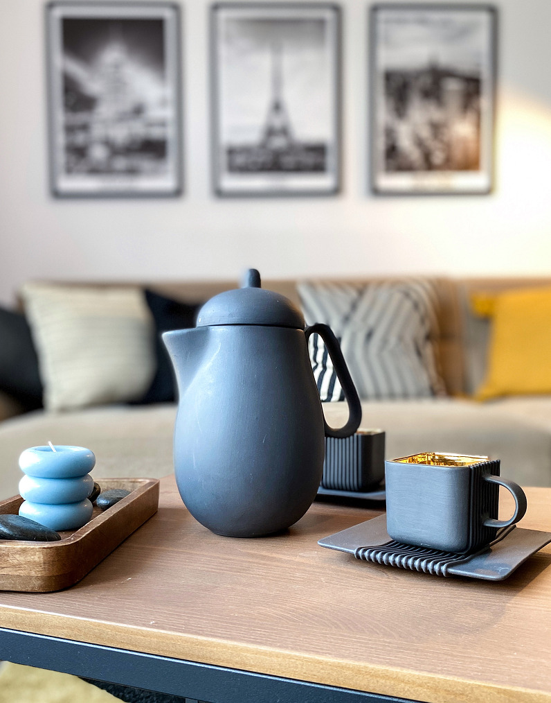 Stylowa aranżacja stolika kawowego z szarą ceramiczną karafką, minimalistyczną filiżanką z złotym wnętrzem, świeczkami w kształcie pączków i czarnymi kamieniami dekoracyjnymi na tle sofy...