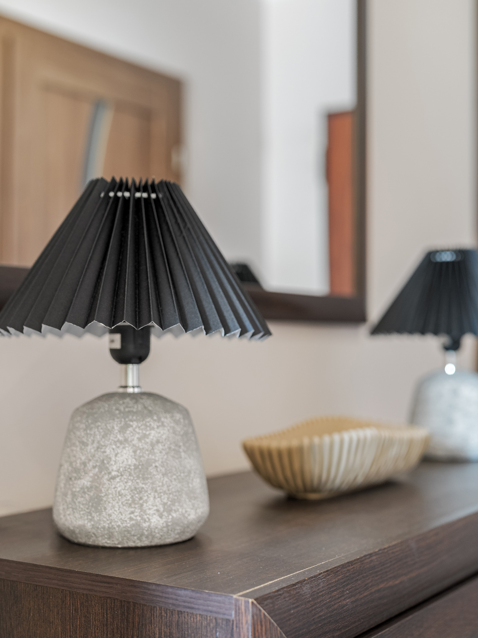 Dwie lampki nocne z czarnymi, plisowanymi abażurami i szarymi, kamiennymi podstawami na ciemnobrązowej komodzie, obok dekoracyjna miseczka w odcieniach beżu.
