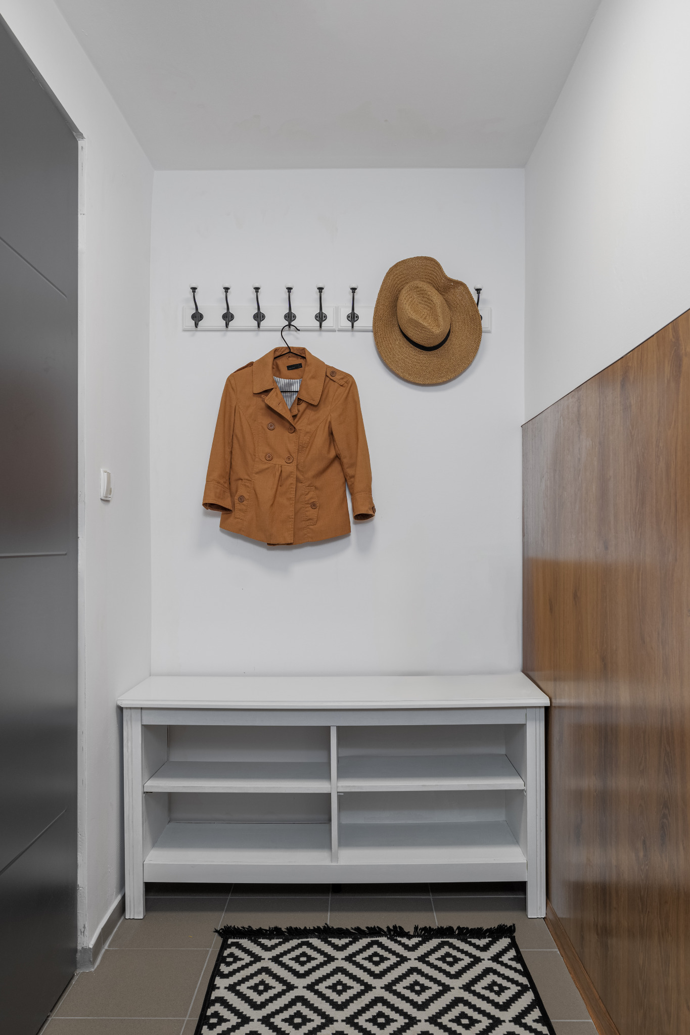 Przedpokój z białymi ścianami, wieszakiem z brązową kurtką i słomkowym kapeluszem, białą szafką na buty i czarno-białym dywanikiem z geometrycznym wzorem.