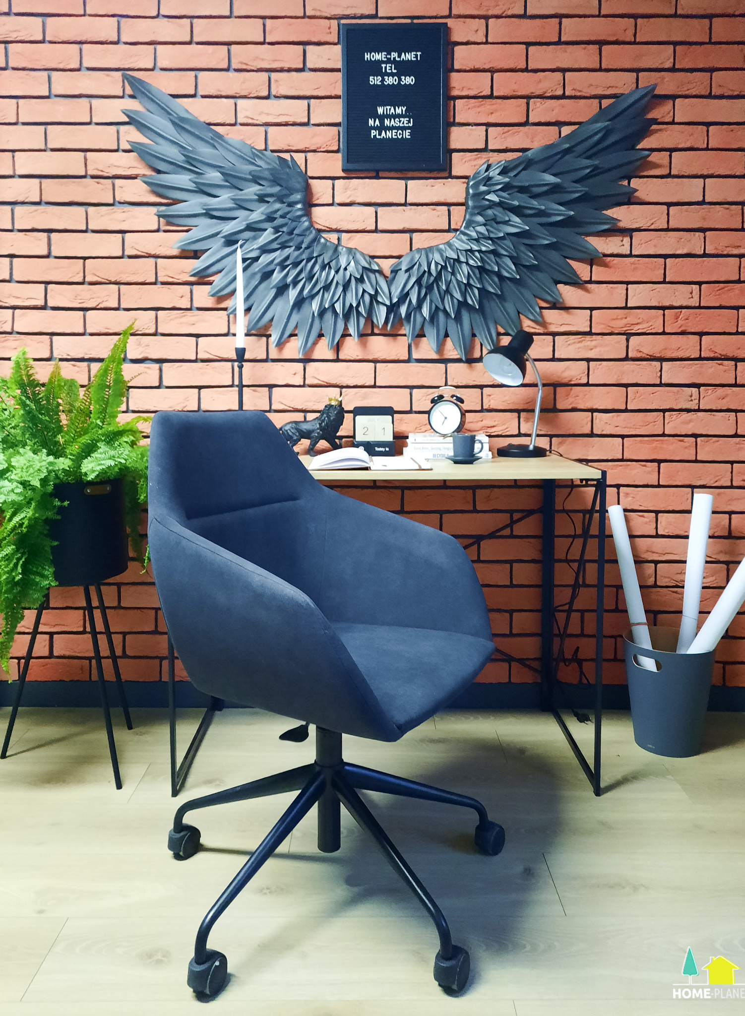 Biuro domowe z szarym krzesłem, biurkiem z lampką, ceglaną ścianą ozdobioną skrzydłami i tablicą z napisem, roślina w doniczce po lewej stronie.