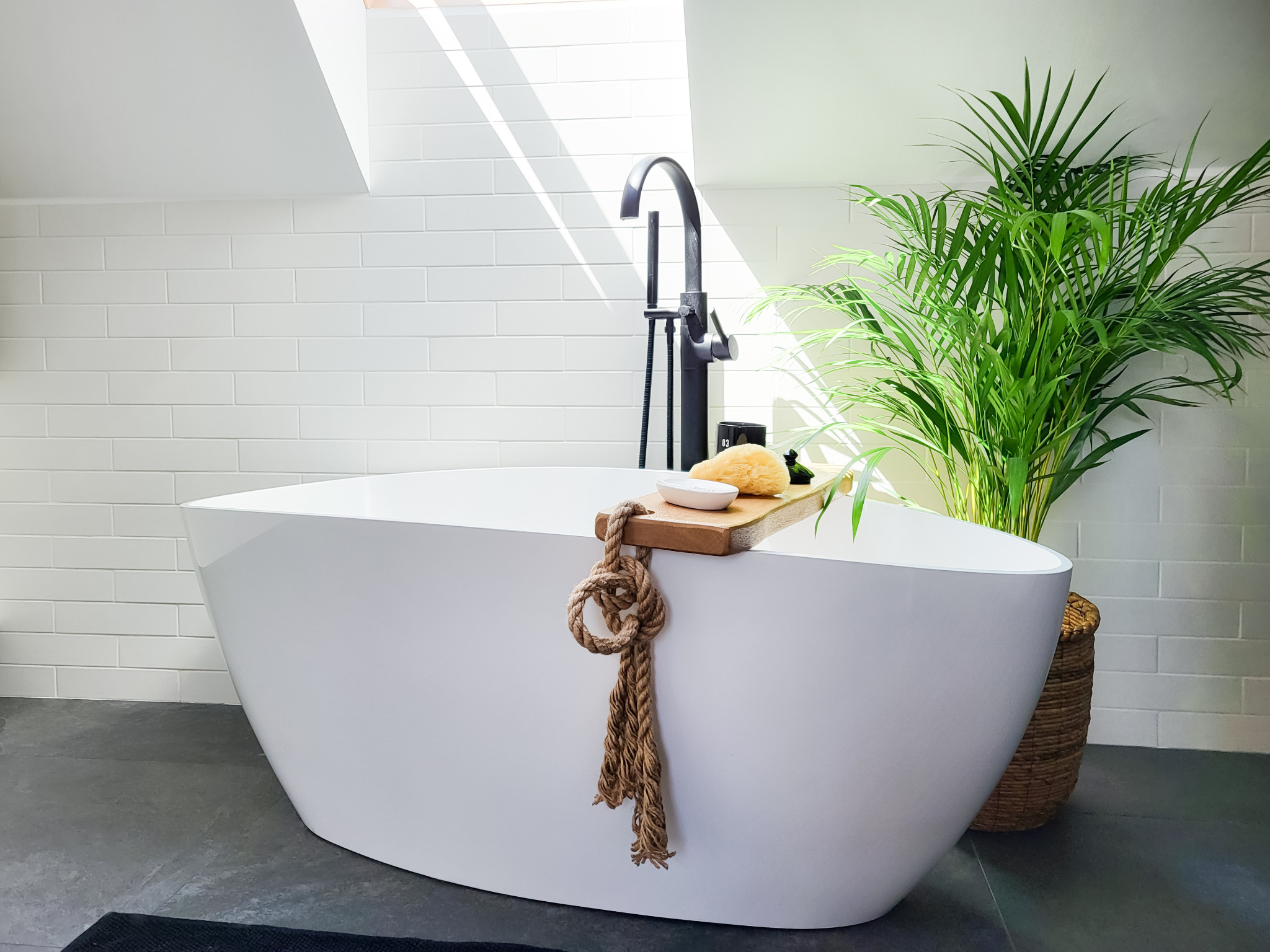 Nowoczesna biała wanna wolnostojąca z czarną armaturą, drewnianą półką z akcesoriami do kąpieli i dekoracyjnym sznurem, ustawiona na ciemnej podłodze w łazience z białymi kafelkami i oknem dachowym...