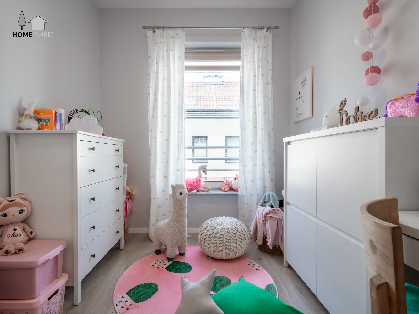 Pokój dziecięcy w jasnych barwach z białymi meblami, różowym dywanem z motywem kaktusów i pluszową lamą. Na parapecie okna siedzą pluszaki, a na komodzie dekoracje.