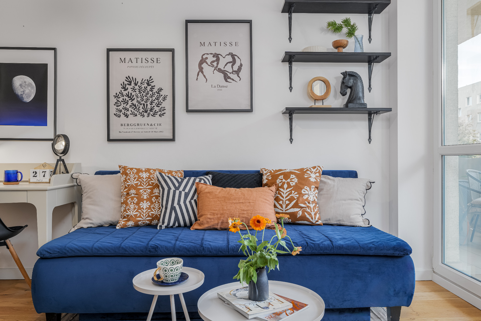 Niebieska sofa z dekoracyjnymi poduszkami i kwiatami w wazonie, stoliki kawowe, grafiki Matisse'a na ścianie, półki z dekoracjami, jasne wnętrze.