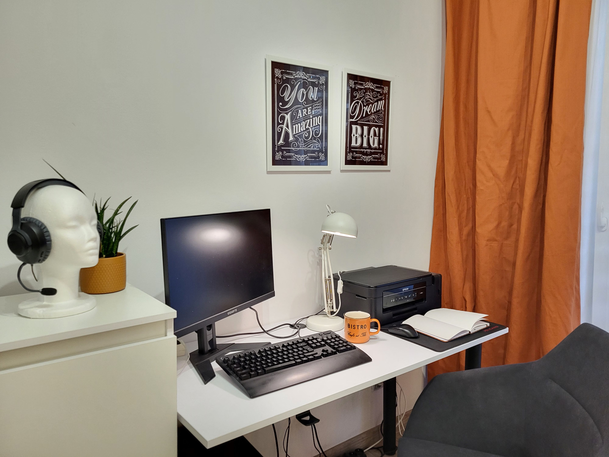 Domowe biuro z białym biurkiem, monitorem, klawiaturą, lampką, drukarką, notatnikiem, pomarańczowym kubkiem Bistro, słuchawkami na manekinie głowy i motywującymi plakatami na ścianie.