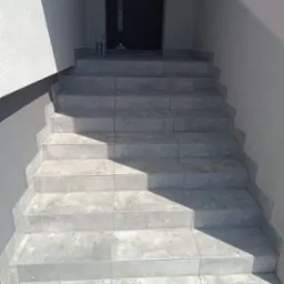 Wykończone płytkami schody wejściowe do budynku, z widocznymi fugami i detalami wykończeniowymi. Na górze dwie tuby silikonu. Częściowo oświetlone światłem słonecznym.