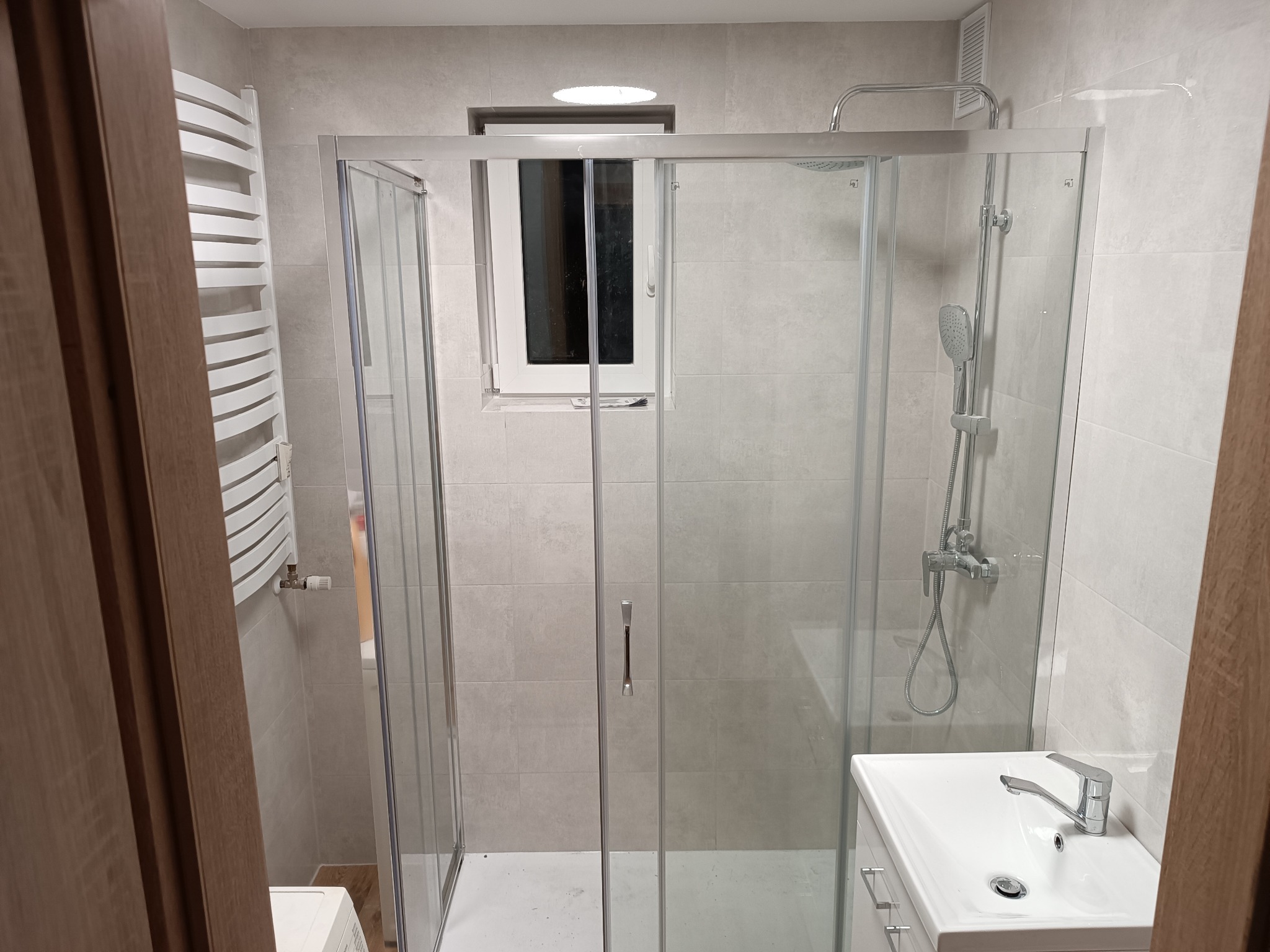 Jasna łazienka po remoncie: kabina prysznicowa ze szklanymi drzwiami, umywalka z szafką, grzejnik drabinkowy i okno. Minimalistyczny styl, płytki w odcieniach szarości.