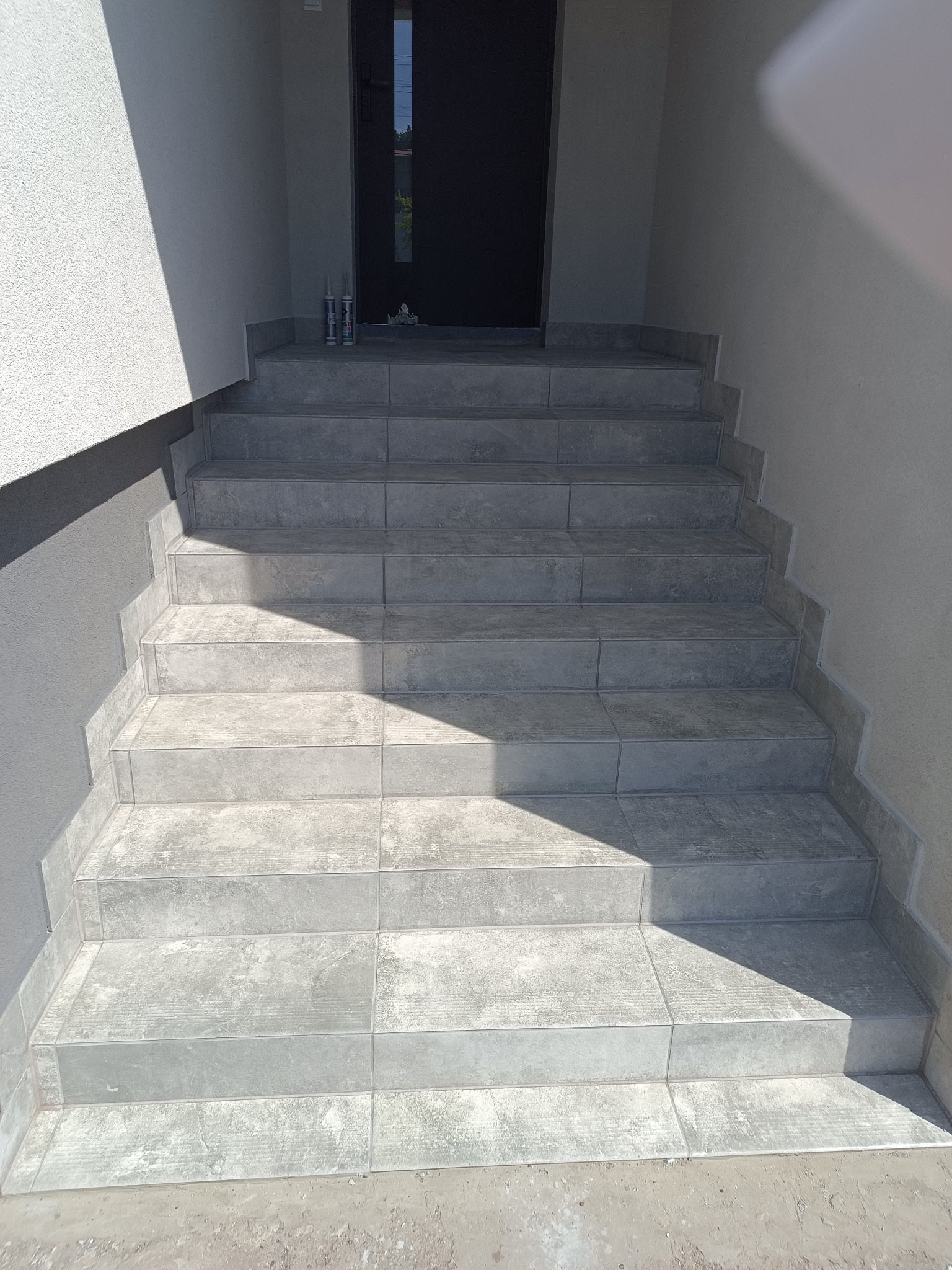 Wykończone płytkami schody wejściowe do budynku, z widocznymi fugami i detalami wykończeniowymi. Na górze dwie tuby silikonu. Częściowo oświetlone światłem słonecznym.