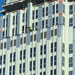 Hedgehog rope access jobs - Pracownicy w odblaskowych kamizelkach pracują na wysokości na elewacji nowoczesnego budynku biurowego z licznymi oknami. Widoczna konstrukcja rusztowania i logo firmy Strabag na szczycie budynku.