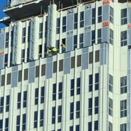 Pracownicy w odblaskowych kamizelkach pracują na wysokości na elewacji nowoczesnego budynku biurowego z licznymi oknami. Widoczna konstrukcja rusztowania i logo firmy Strabag na szczycie budynku.