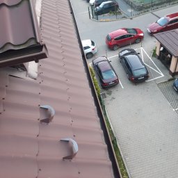Hedgehog rope access jobs - Widok z góry na brązowy dach z blachy trapezowej z widocznymi uchwytami rynnowymi, na tle parkingu z samochodami i budynkiem.
