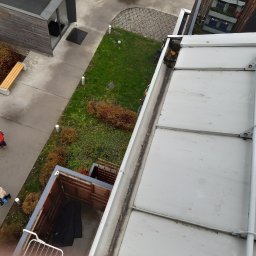Hedgehog rope access jobs - Widok z góry na zabrudzoną rynnę dachową, z widocznymi liśćmi i osadami, na tle fragmentu budynku z balkonem i zielenią miejską.