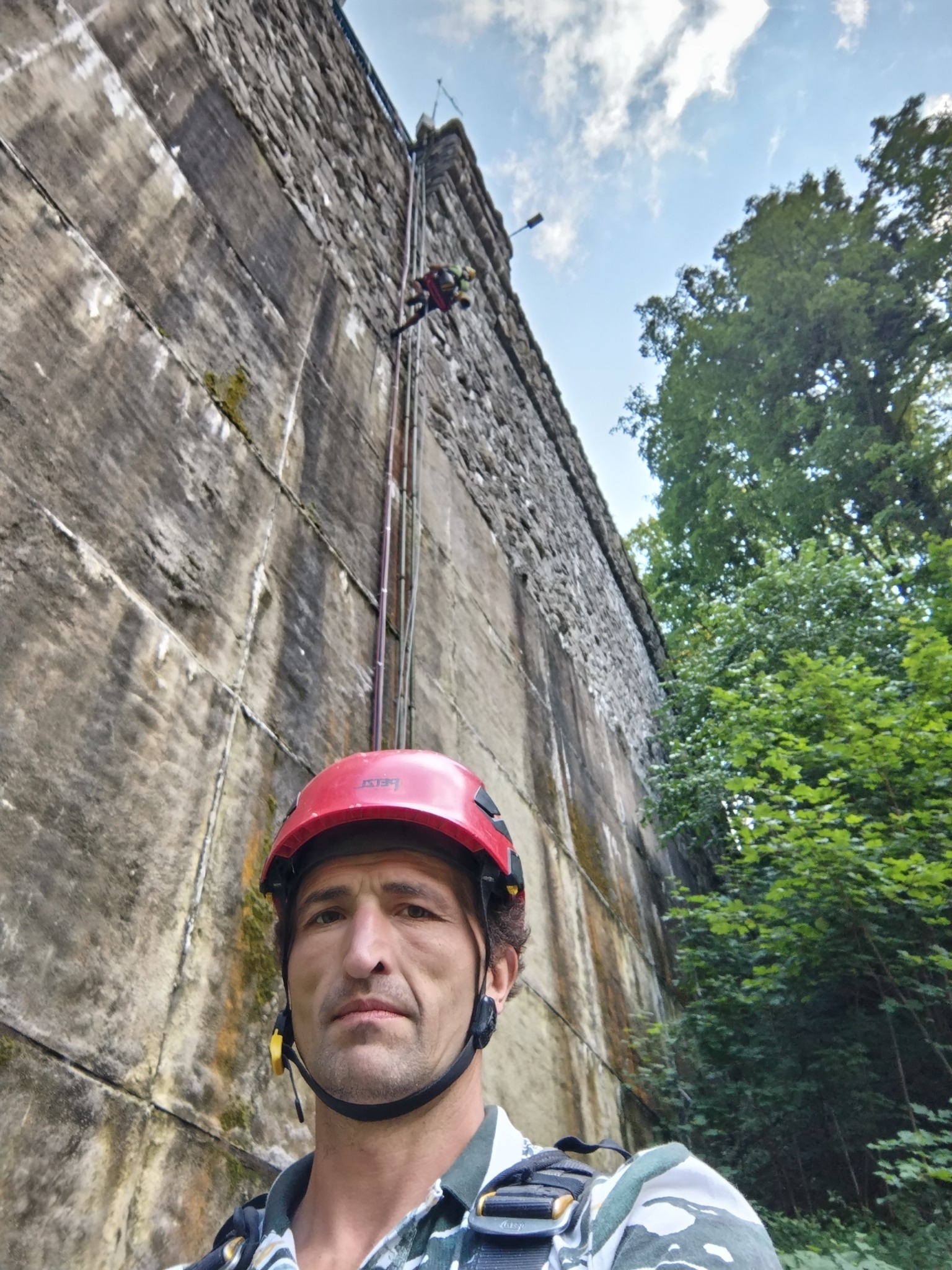 Mężczyzna w kasku, z widoczną uprzężą, stoi przed wysoką, kamienną ścianą; w tle drugi pracownik na linie, prawdopodobnie podczas prac na wysokości.
