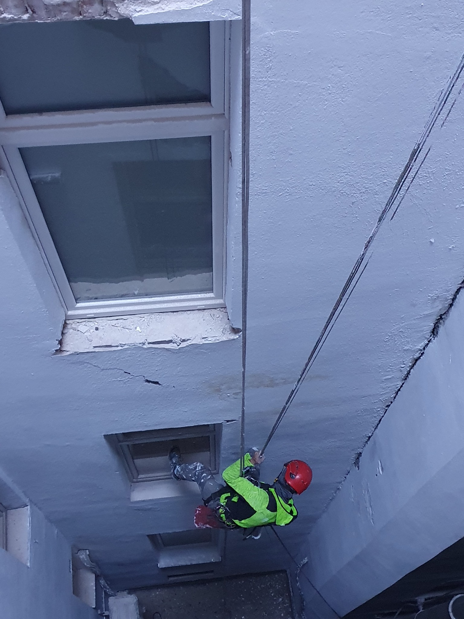 Pracownik na linach w kasku i kamizelce asekuracyjnej podczas pracy na wysokości przy budynku z oknami i widocznymi uszkodzeniami elewacji.