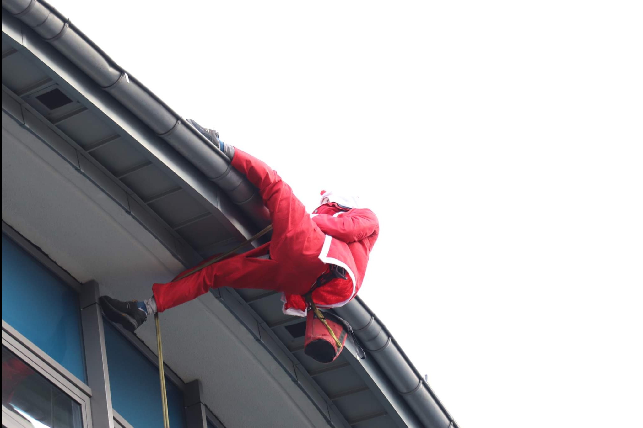 Osoba przebrana za Świętego Mikołaja, na linie, zwisa z rynny budynku podczas potencjalnego czyszczenia. Widoczny kubeł zawieszony na linie.