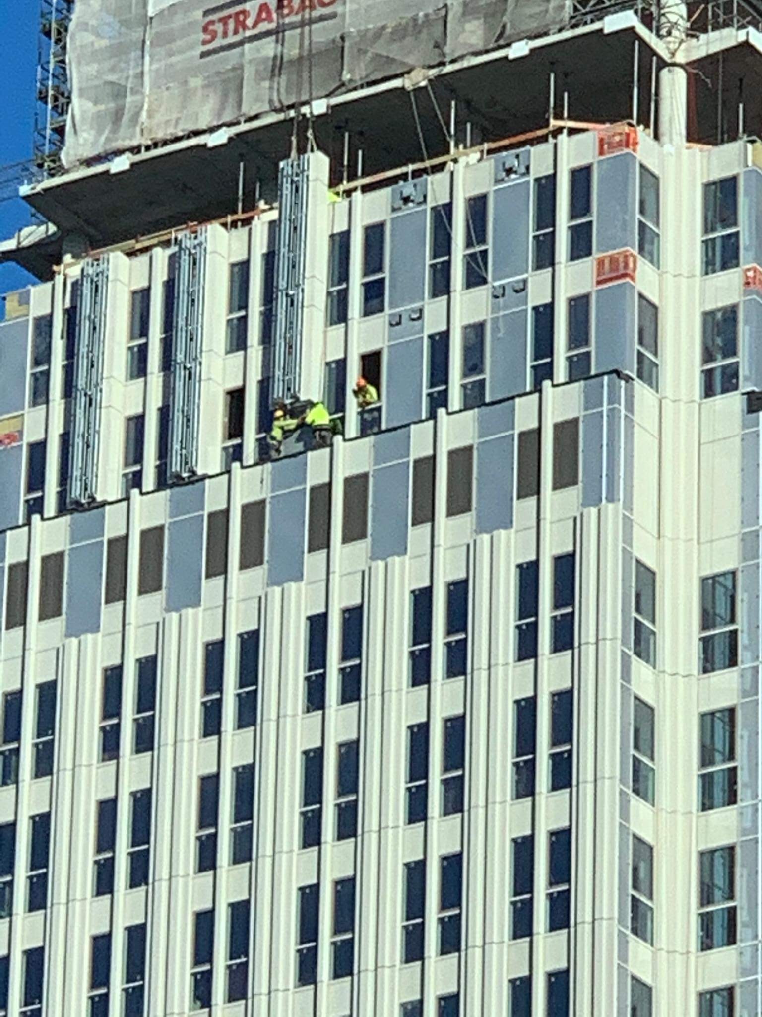 Pracownicy w odblaskowych kamizelkach pracują na wysokości na elewacji nowoczesnego budynku biurowego z licznymi oknami. Widoczna konstrukcja rusztowania i logo firmy Strabag na szczycie budynku.