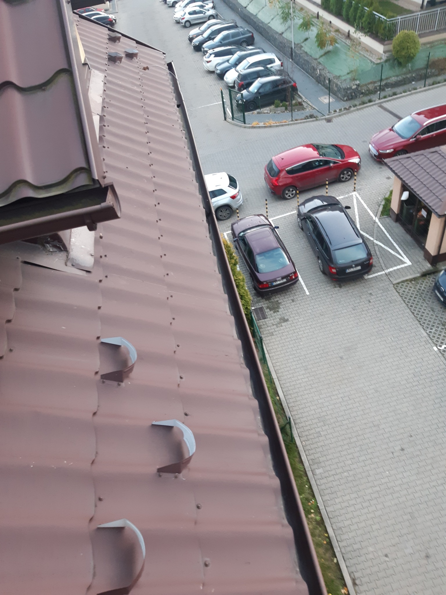 Widok z góry na brązowy dach z blachy trapezowej z widocznymi uchwytami rynnowymi, na tle parkingu z samochodami i budynkiem.