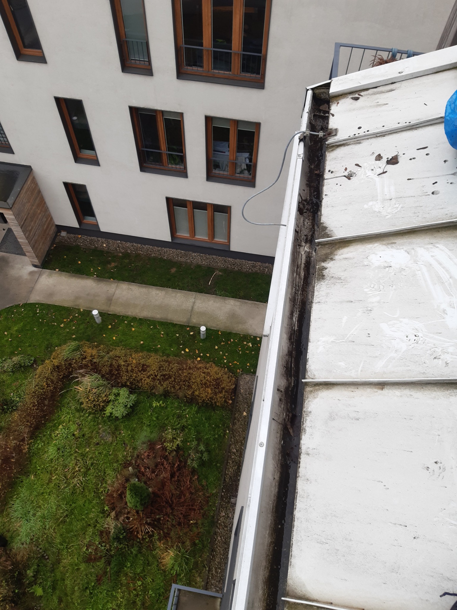 Widok z góry na brudną rynnę dachową wzdłuż krawędzi dachu budynku mieszkalnego, z widocznym podwórkiem z zielenią i chodnikiem.