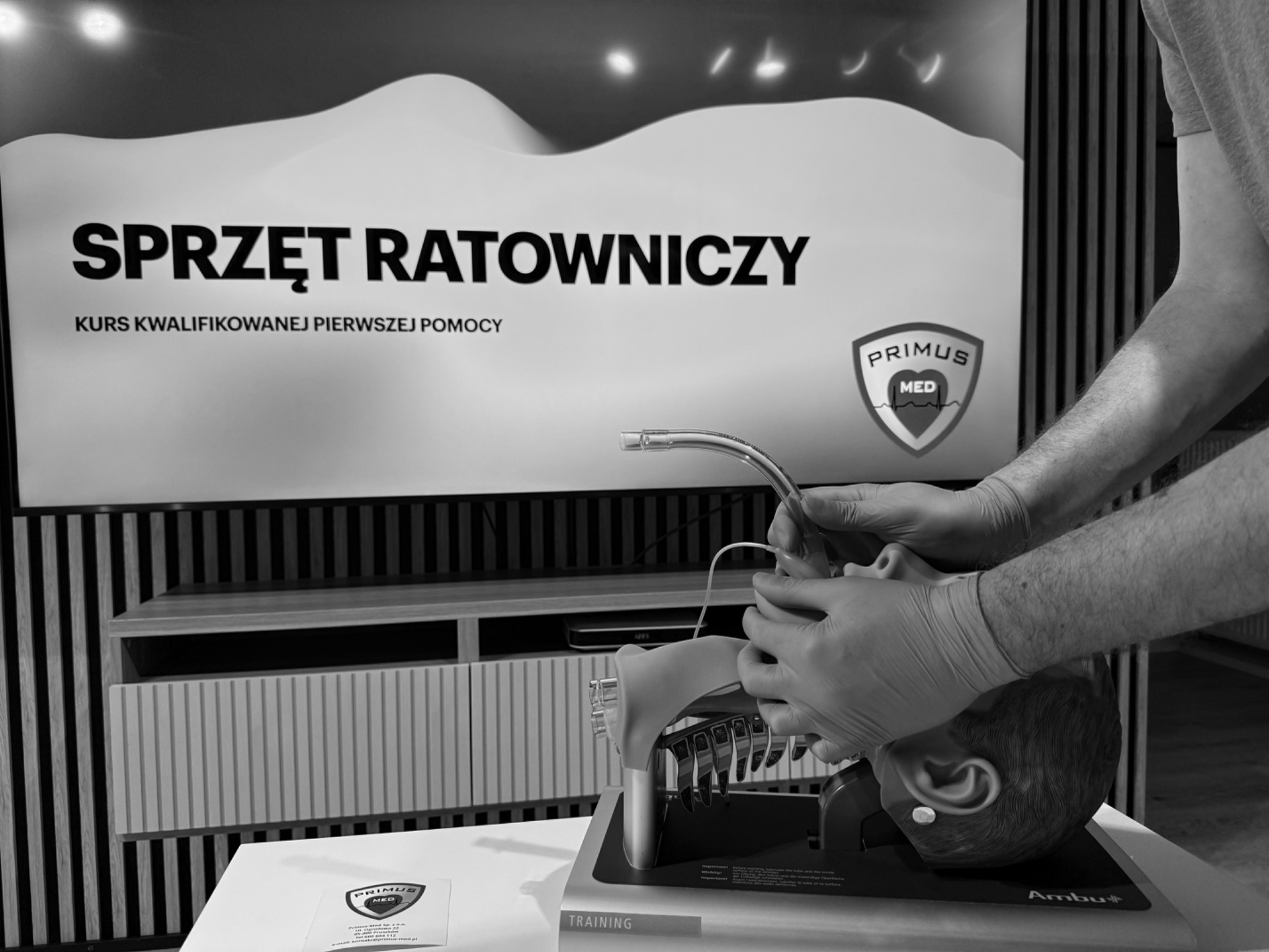 Instruktor w rękawiczkach demonstruje intubację na fantomie podczas kursu kwalifikowanej pierwszej pomocy; w tle baner z napisem 'Sprzęt Ratowniczy' i logo Primus Med.