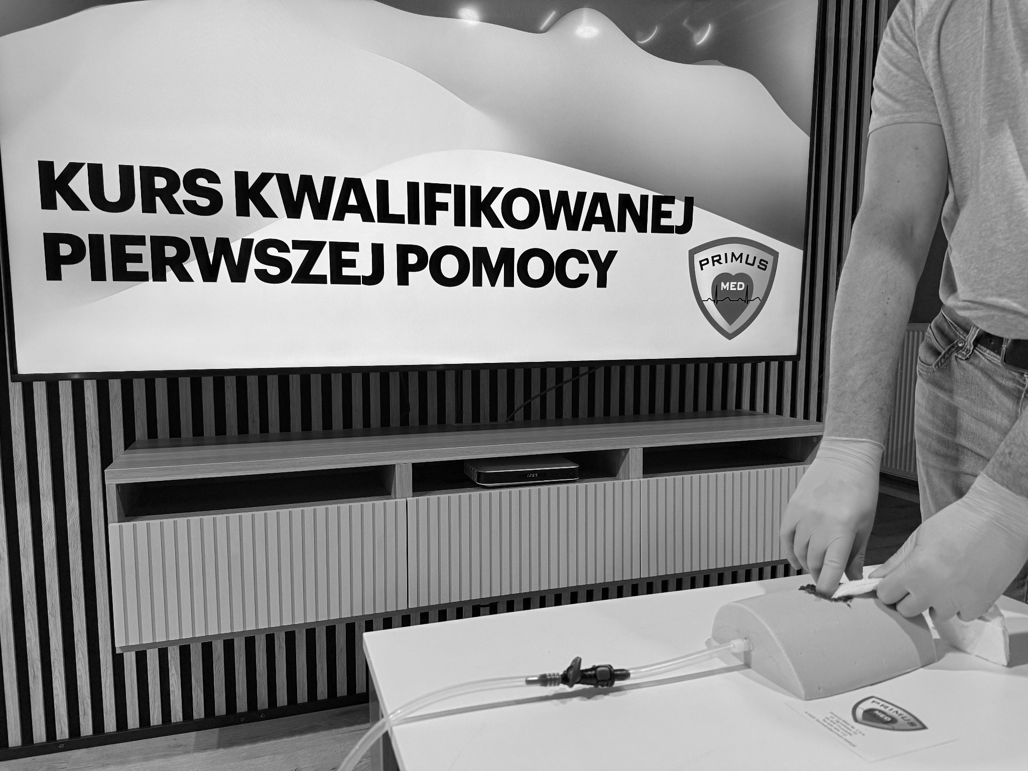 Instruktor w rękawicach lateksowych demonstruje opatrywanie rany na fantomie podczas kursu kwalifikowanej pierwszej pomocy, w tle ekran z napisem 'Kurs Kwalifikowanej Pierwszej Pomocy' i logo...