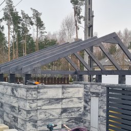 Złota rączka Mateusz Biały - Konstrukcja z ciemnego drewna na murowanym słupku ogrodzenia z szarej kostki, widoczne narzędzia i rękawice robocze, w tle dom i drzewa.